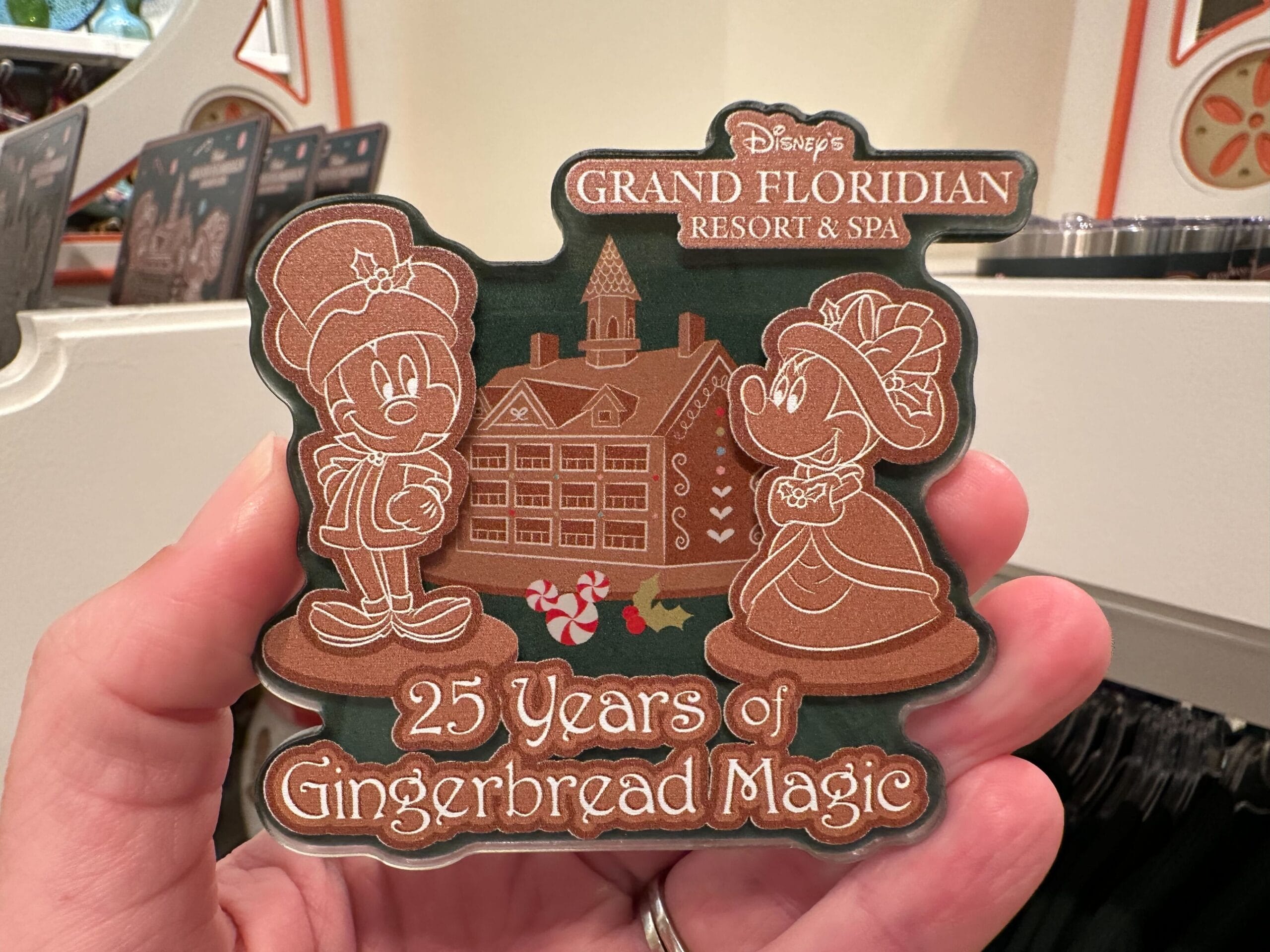 A hand holds a souvenir featuring cartoon characters celebrating 25 Years of Gingerbread Magic at Disney's Grand Floridian Resort & Spa against a festive background.