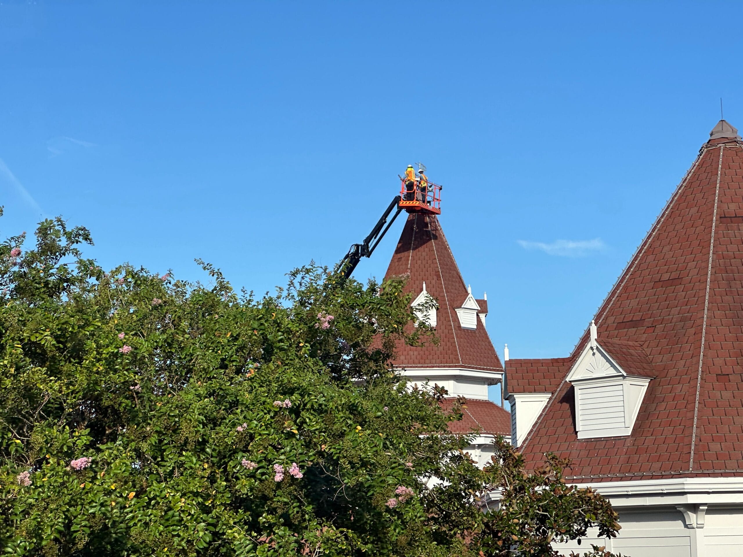 Workers in a lift repair the pointed roof of a Grand Floridian building next to trees under a clear blue sky.