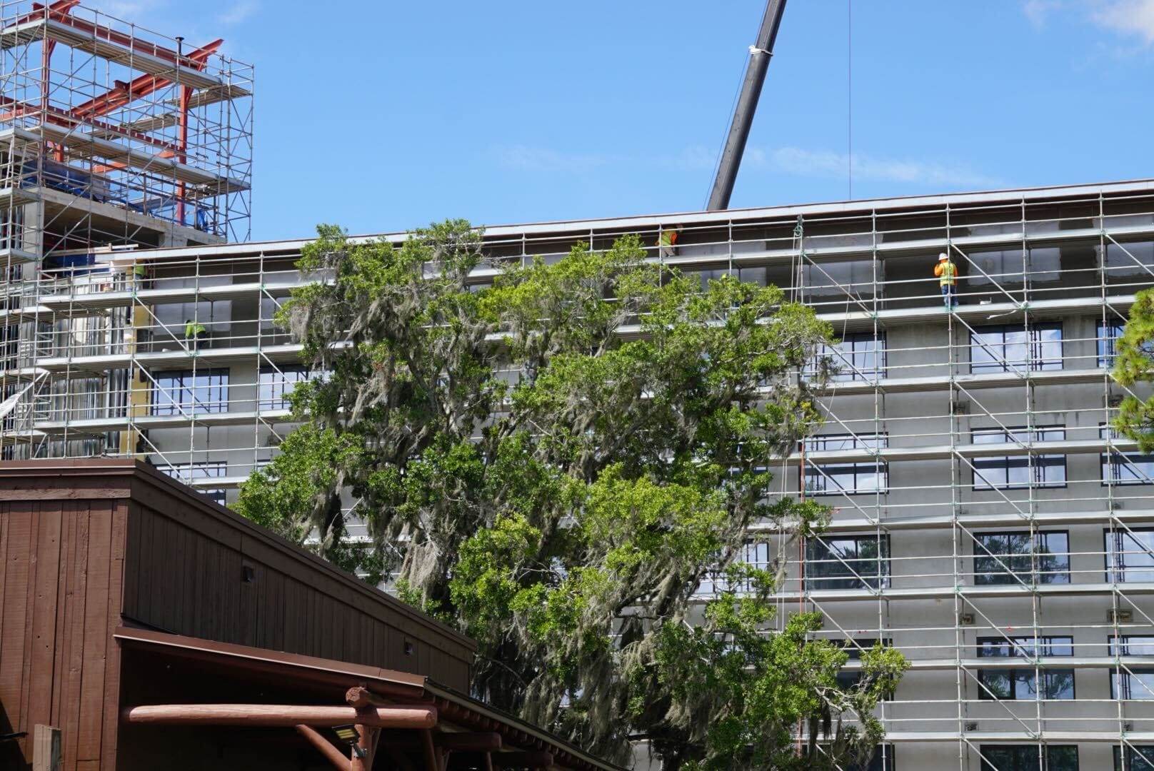 Workers on scaffolding renovate a building behind trees and a wooden structure under a clear blue sky.