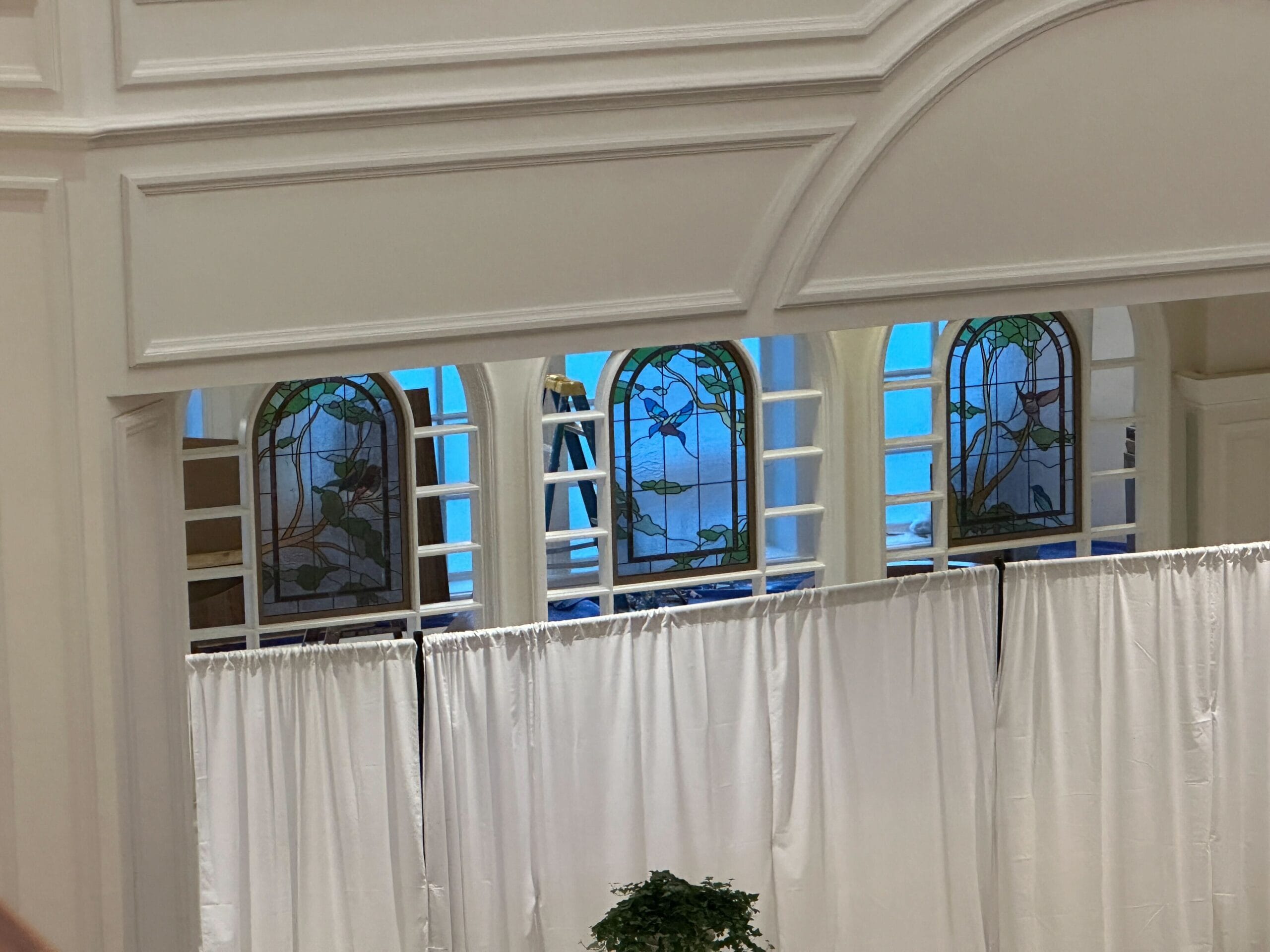 A ladder stands behind white curtains in front of Tea Room Windows inside a white building.