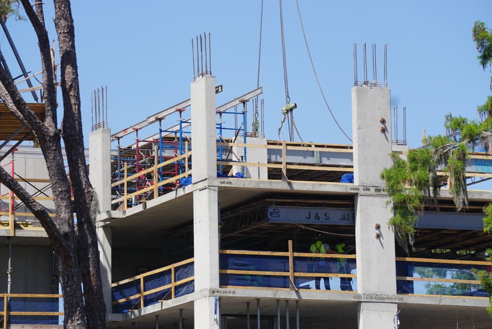 A construction update reveals workers on a multi-story building, with concrete columns and metal frames prominently displayed. A crane lifts materials against the backdrop of trees, as they enhance the progress of the future Lakeshore Lodge.