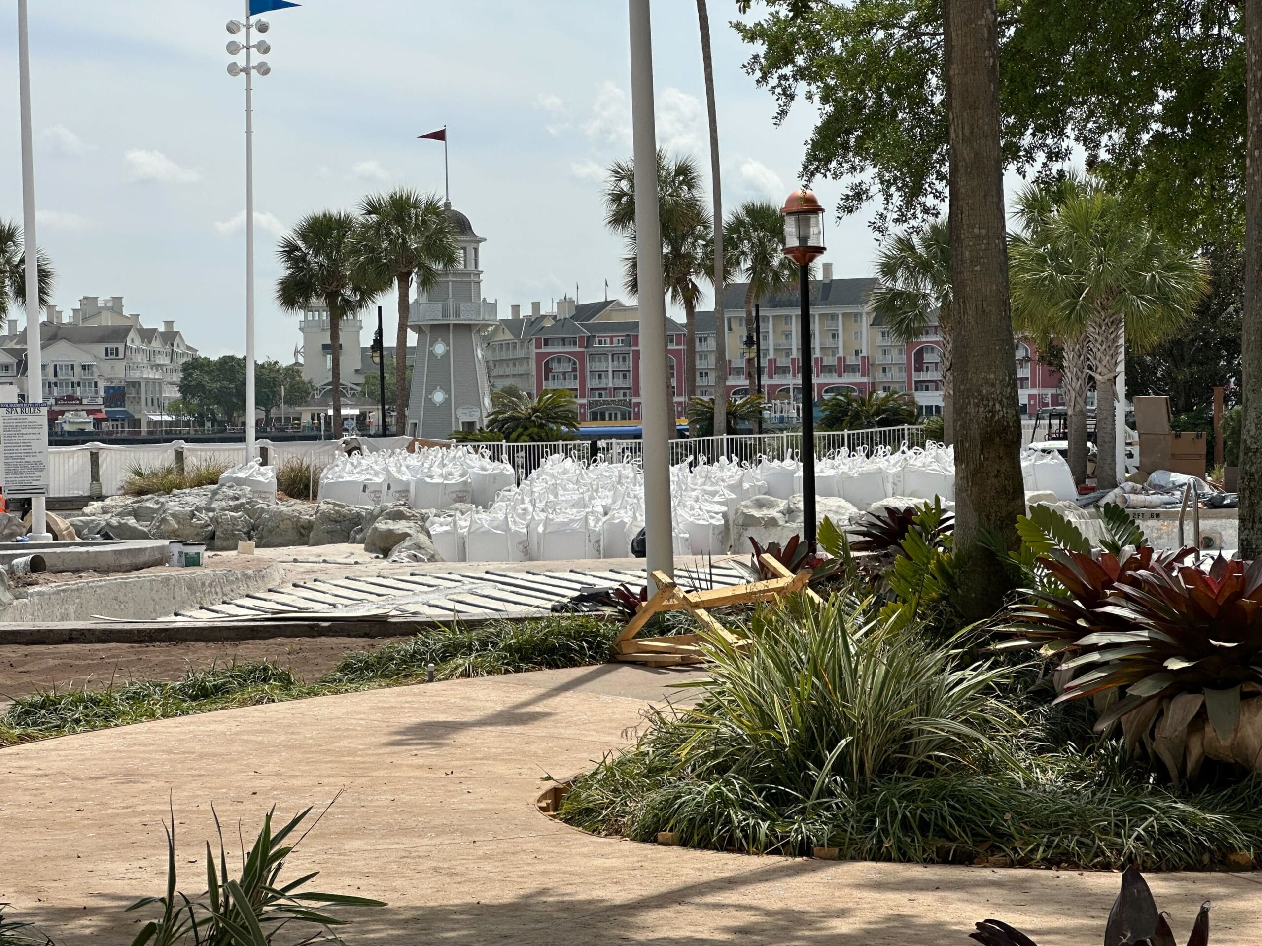 Construction site with scattered materials like white blocks and wooden planks near a path in a garden setting, hinting at pool construction. In the background, buildings and palm trees stand under a cloudy sky, reminiscent of Disney's Yacht Club ambiance.