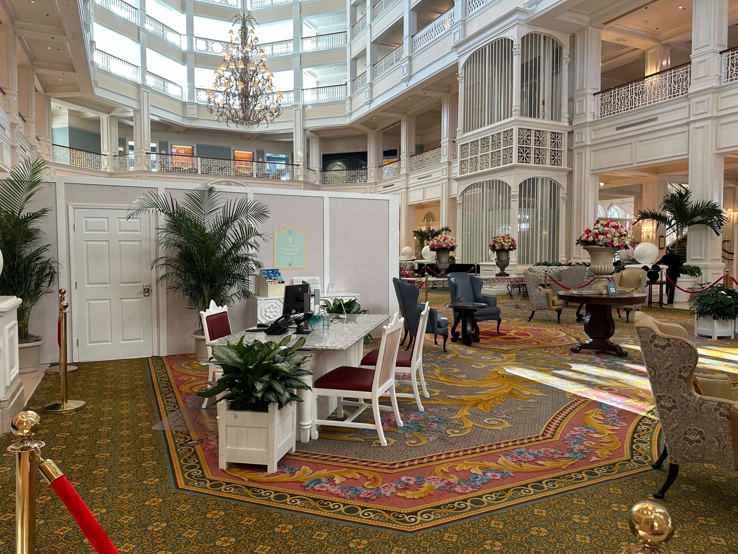Elegant hotel lobby with high ceilings, chandelier, marble desk, chairs, plants, and ornate carpet.