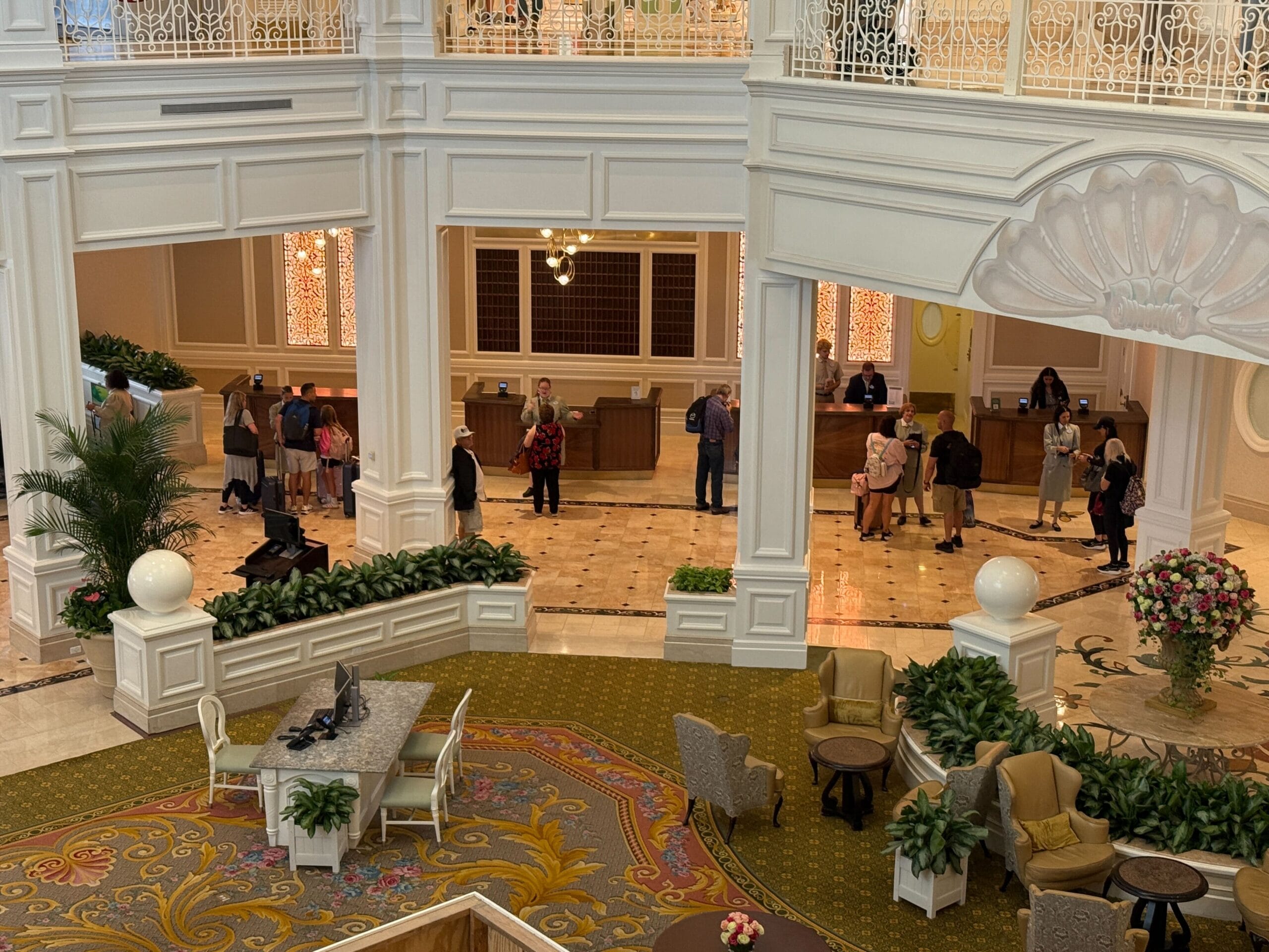 Hotel lobby with people waiting in line at the reception desk and a seating area with chairs, tables, and plants.