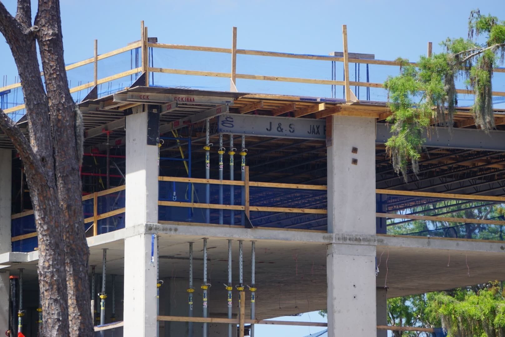 Partially constructed building with concrete pillars, wooden railings, and temporary supports. Blue tarp is draped over sections. Trees partially visible in the foreground.