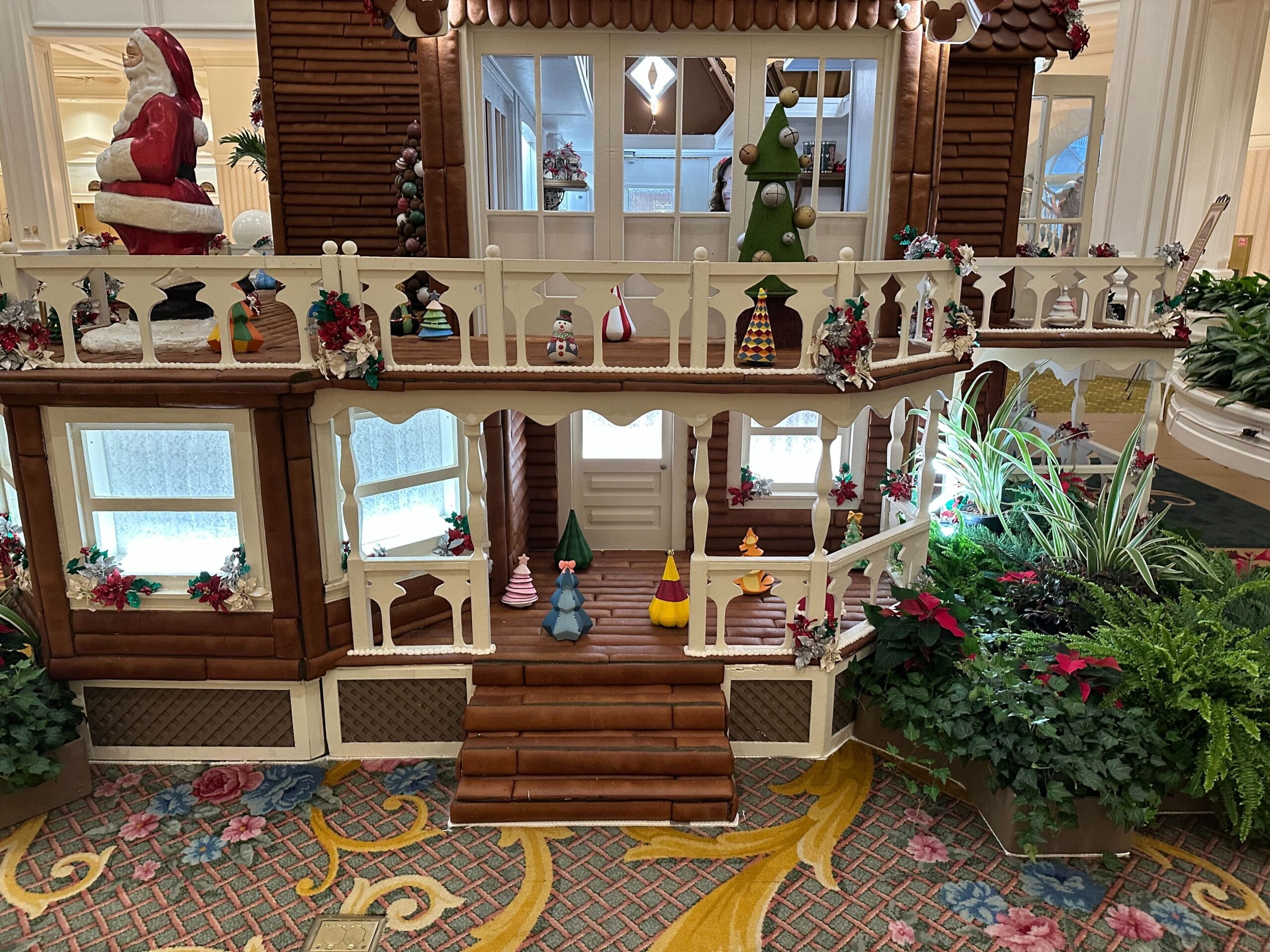 A festive gingerbread house display with mini holiday figures and decorations, surrounded by green plants and red flowers.