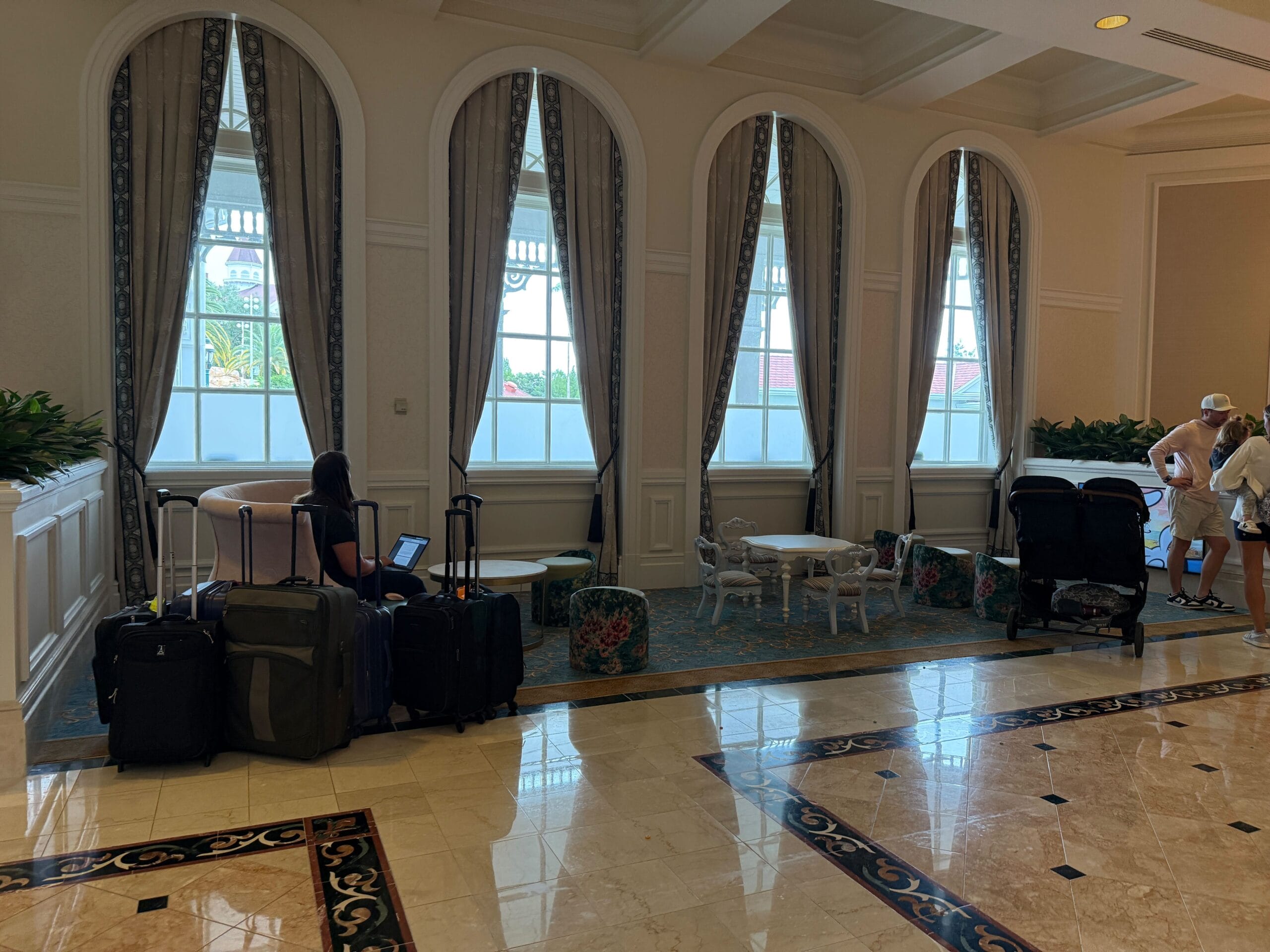 A woman with luggage sits in a hotel lobby near large windows; people and strollers are on the right.