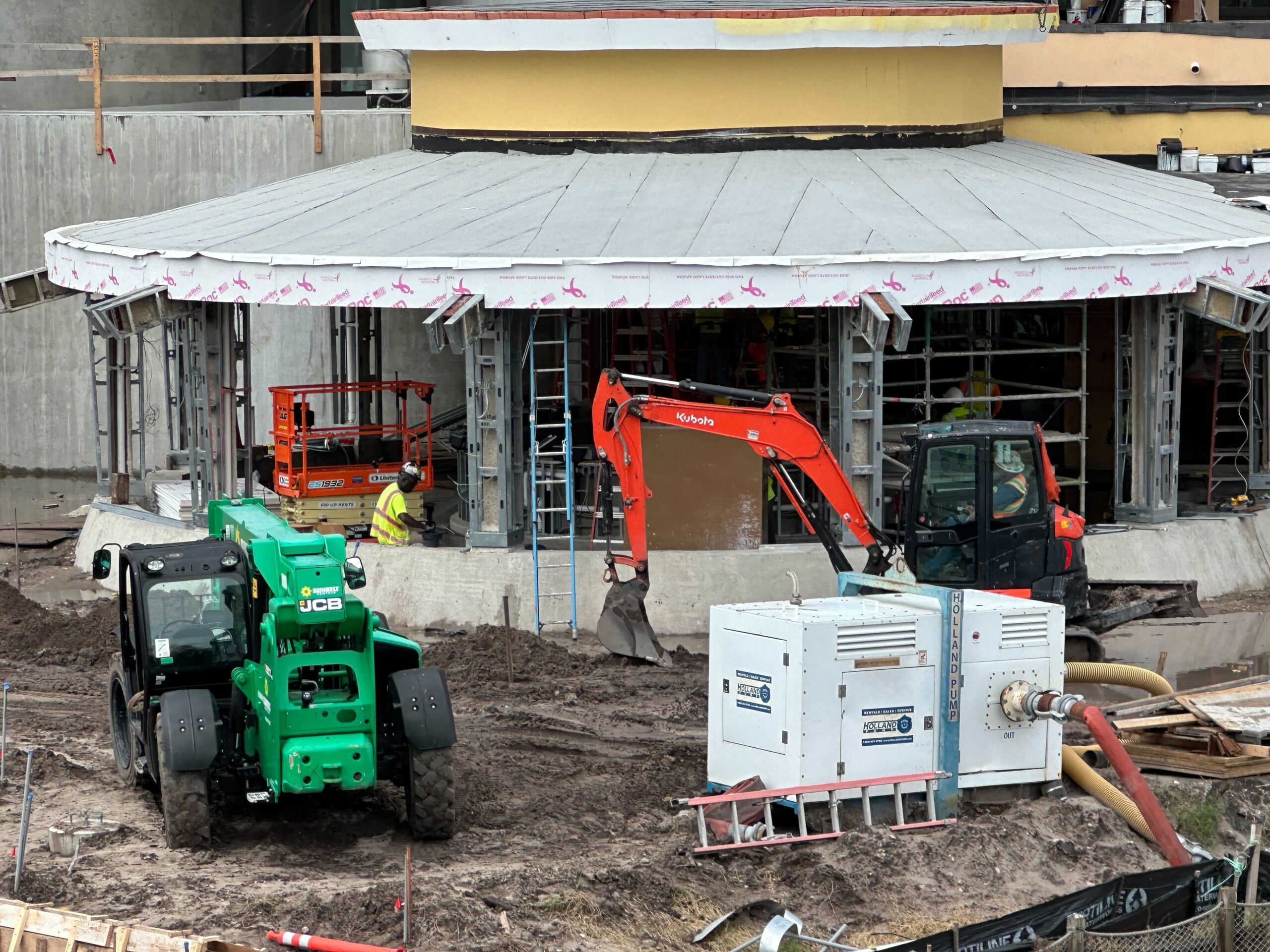 Construction site with workers and machinery, including a green telehandler, an orange excavator, a red compact loader, and an industrial unit, in front of a building under construction.