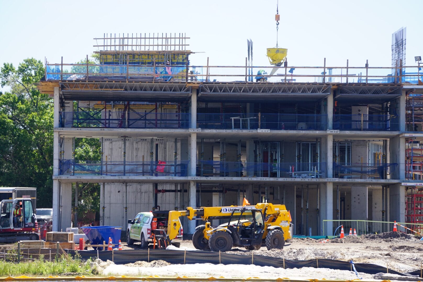 Construction site with a partially built multi-story building. Workers, a crane, and heavy machinery, including a yellow forklift, are visible against a clear sky.