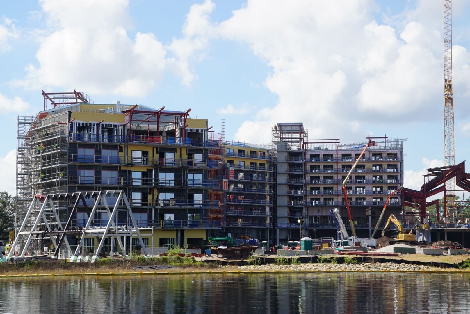 A multi-story Disney Lakeshore Lodge under construction with cranes and scaffolding beside a body of water on a partly cloudy day.