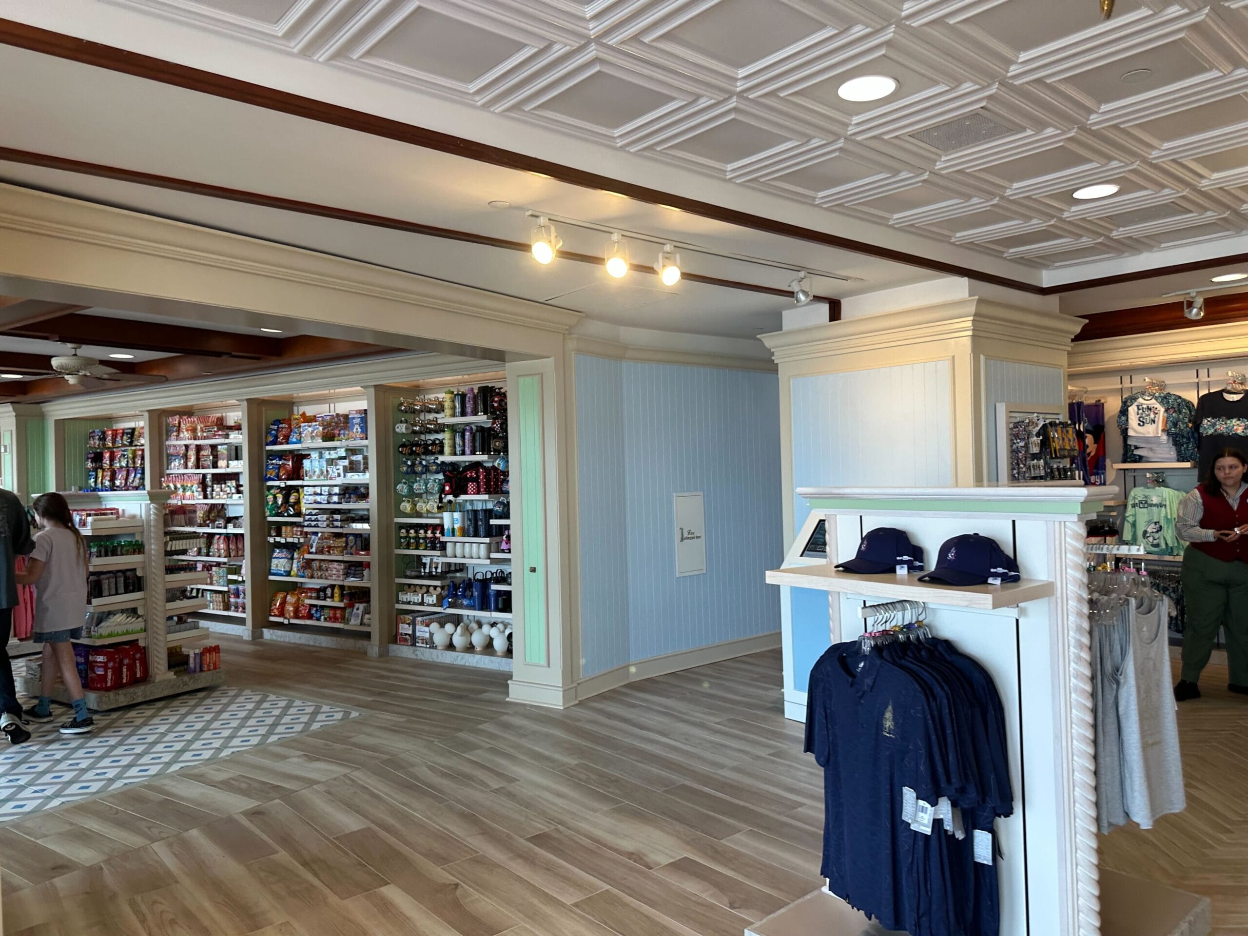 A store interior with wooden flooring at Disney's Boardwalk Inn showcases shelves of snacks, drinks, and clothing. A stand featuring folded shirts and hats sits prominently in the renovated section. Two people are browsing contentedly.