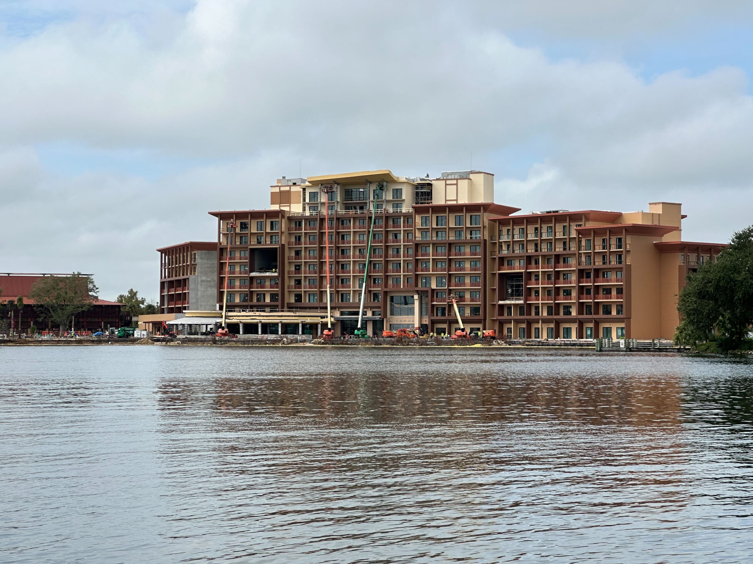 A large multistory building under construction is seen across a body of water with a crane and various construction equipment on site. The sky is partly cloudy.