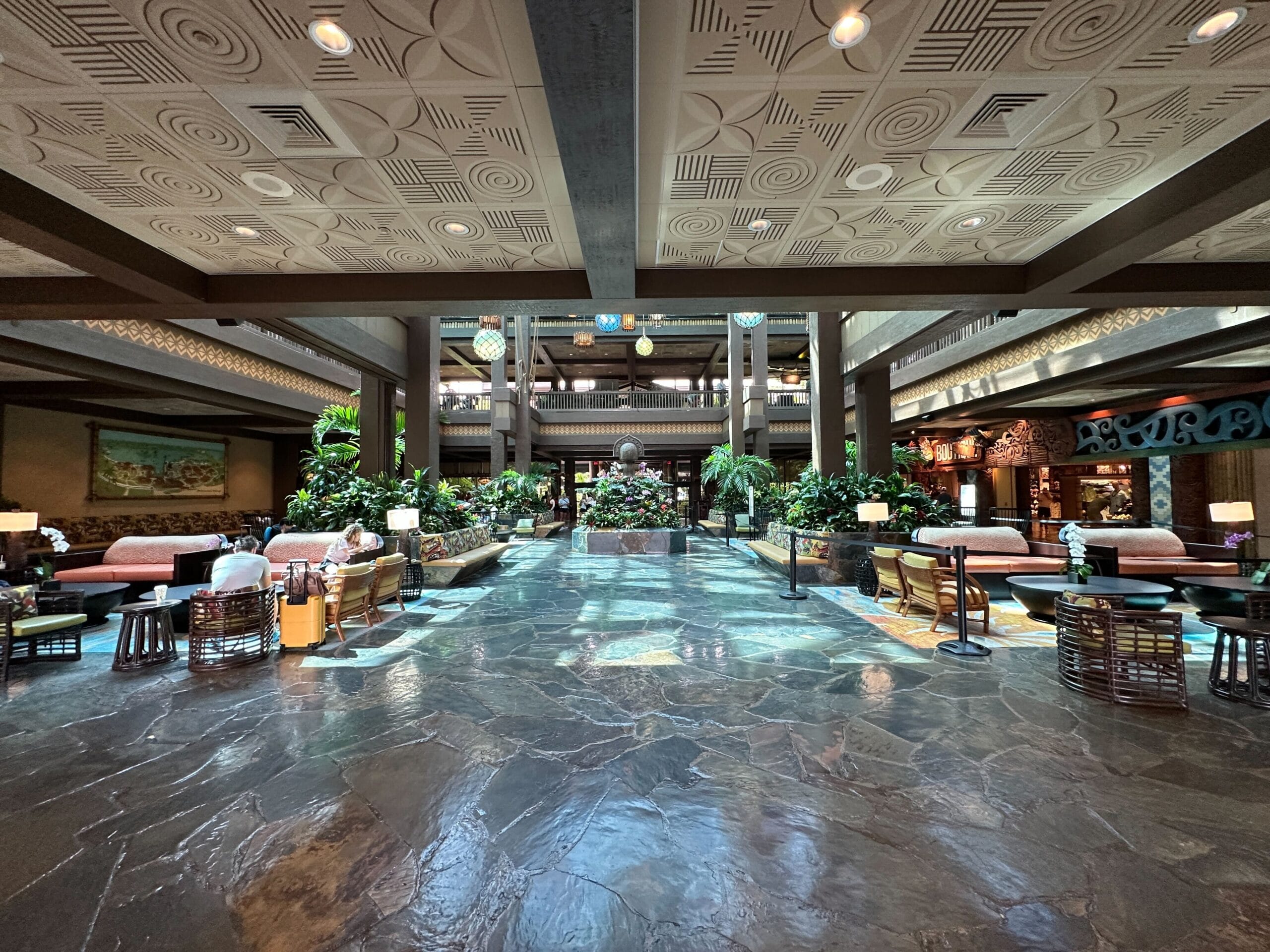 Spacious hotel lobby with tropical decor, stone flooring, lush plants, and seating areas on both sides.