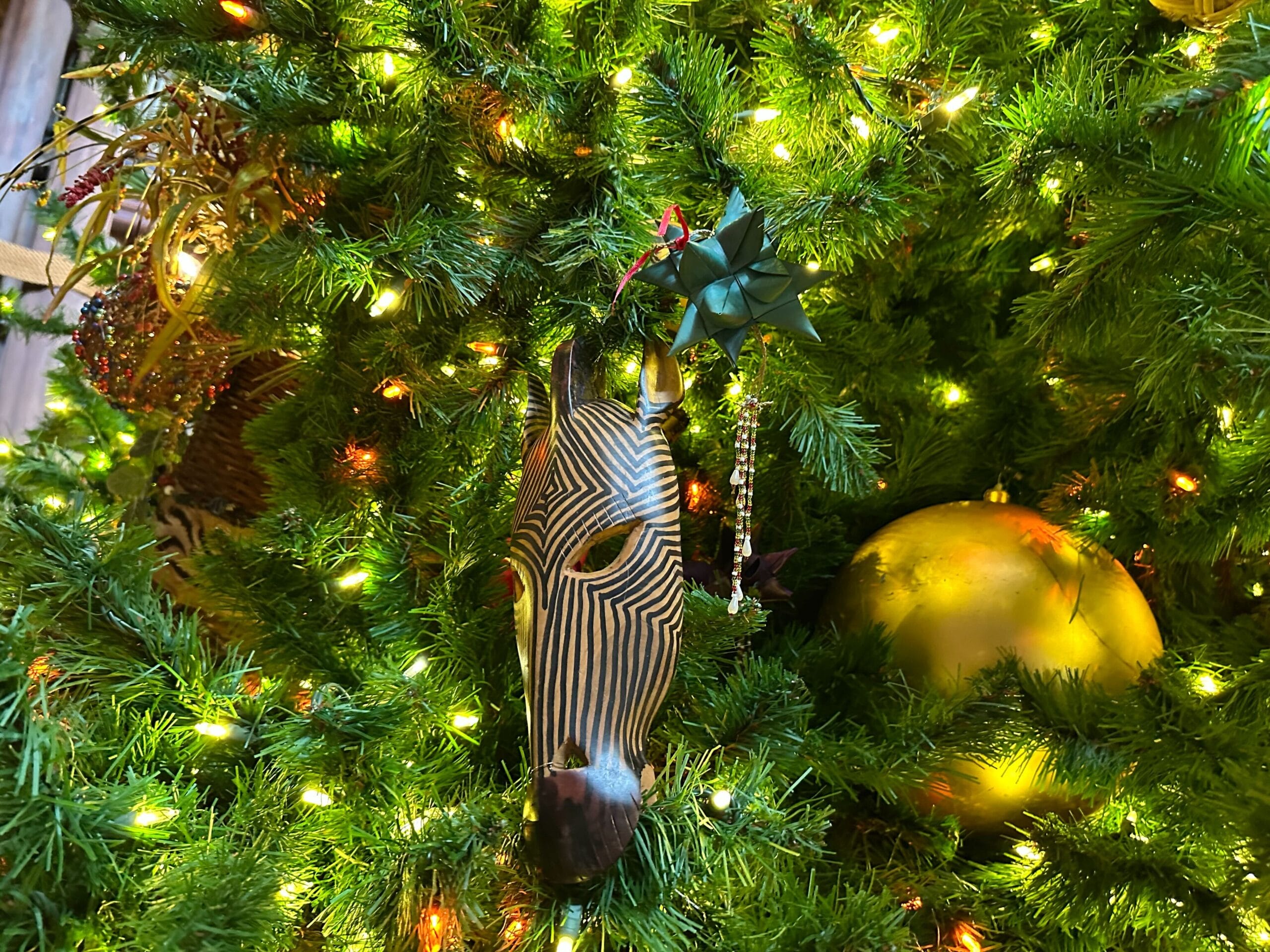 A zebra mask hangs on a Christmas tree adorned with lights, a gold ornament, and a green bow.