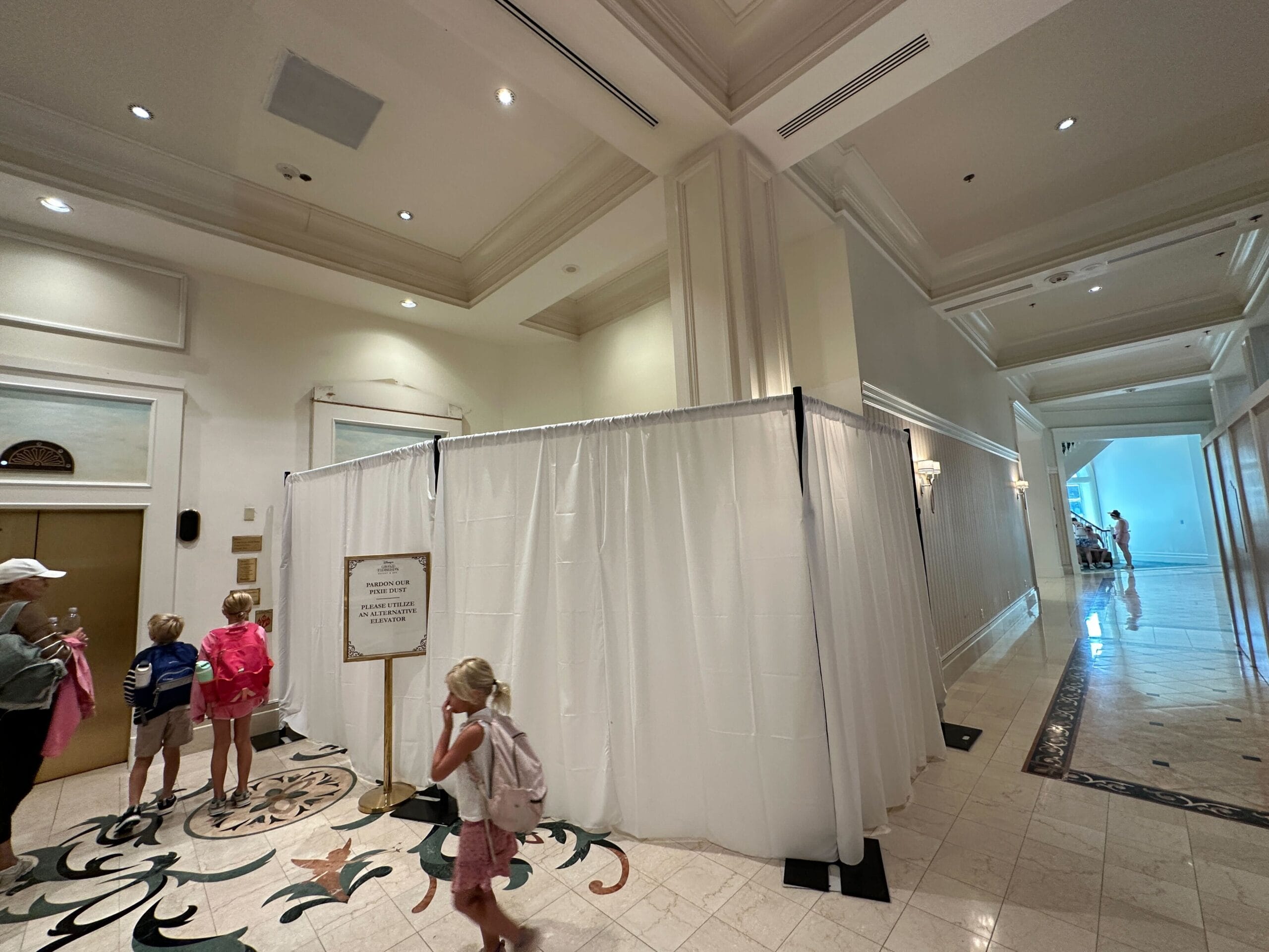 Children and adults walk past curtained-off Grand Floridian Construction in a hotel lobby with tiled floors and high ceilings.