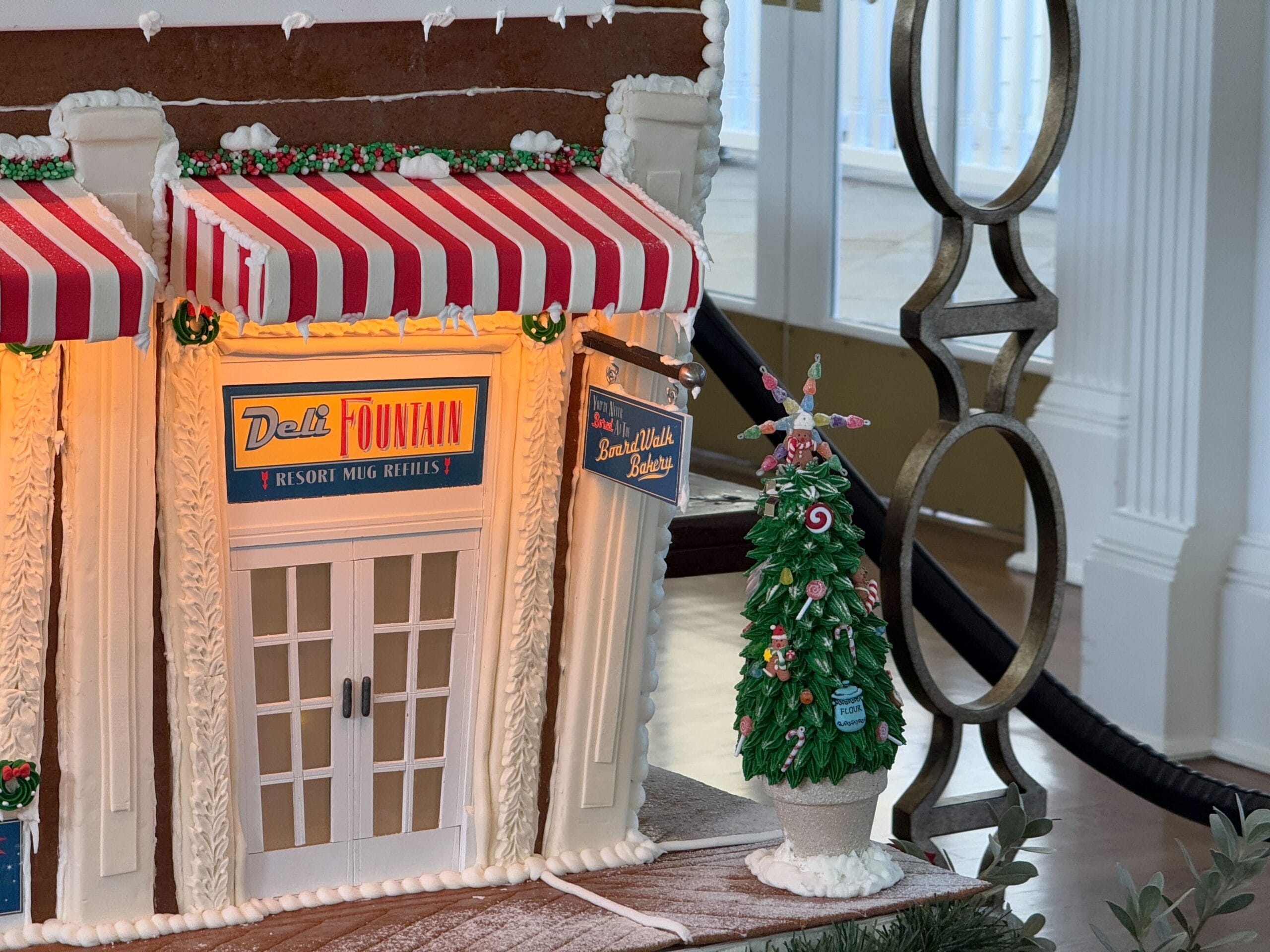 Gingerbread house styled as a deli, with striped awnings and a small decorated Christmas tree beside it.