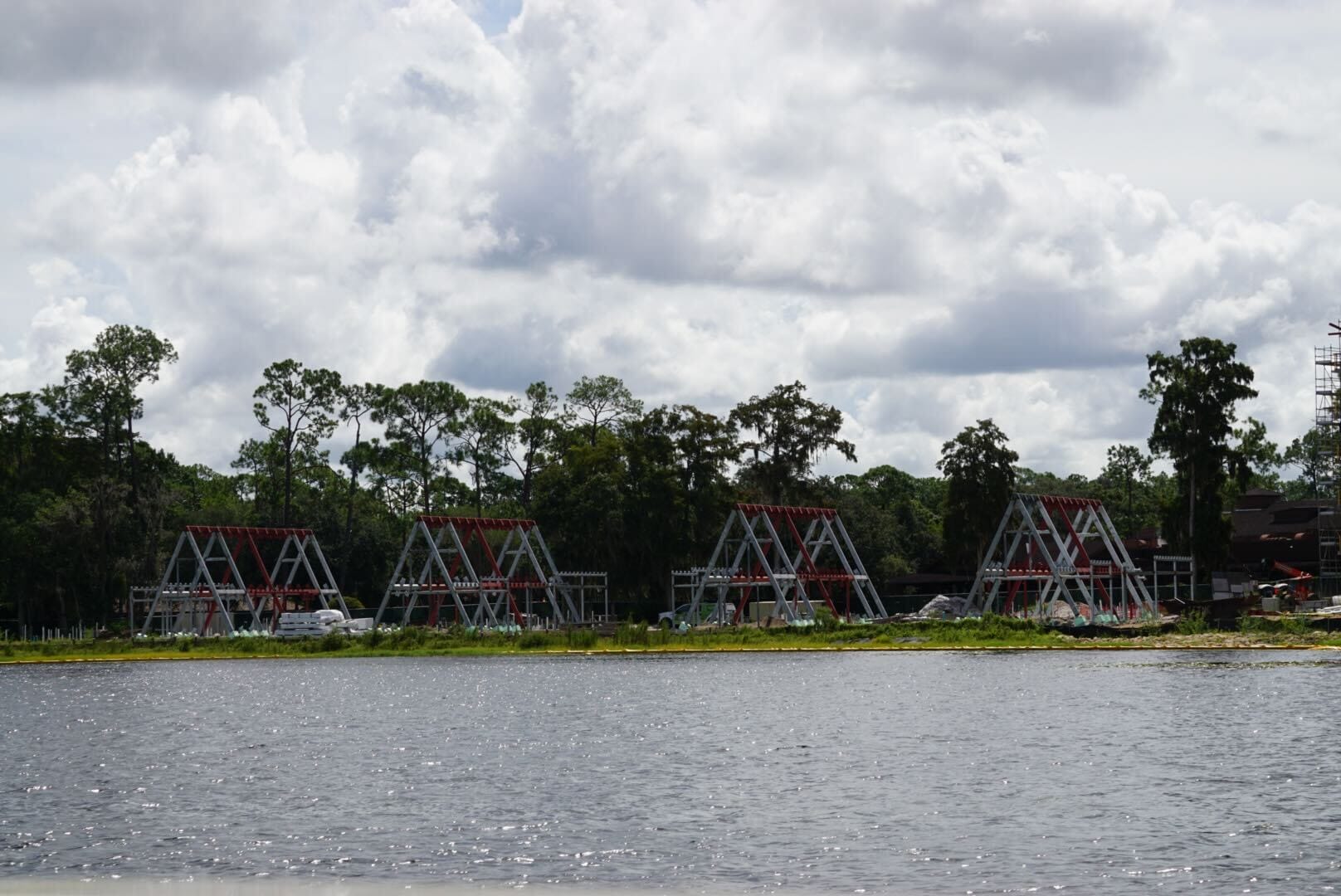 Four triangular metal structures under construction by a lake, with trees and clouds in the background.