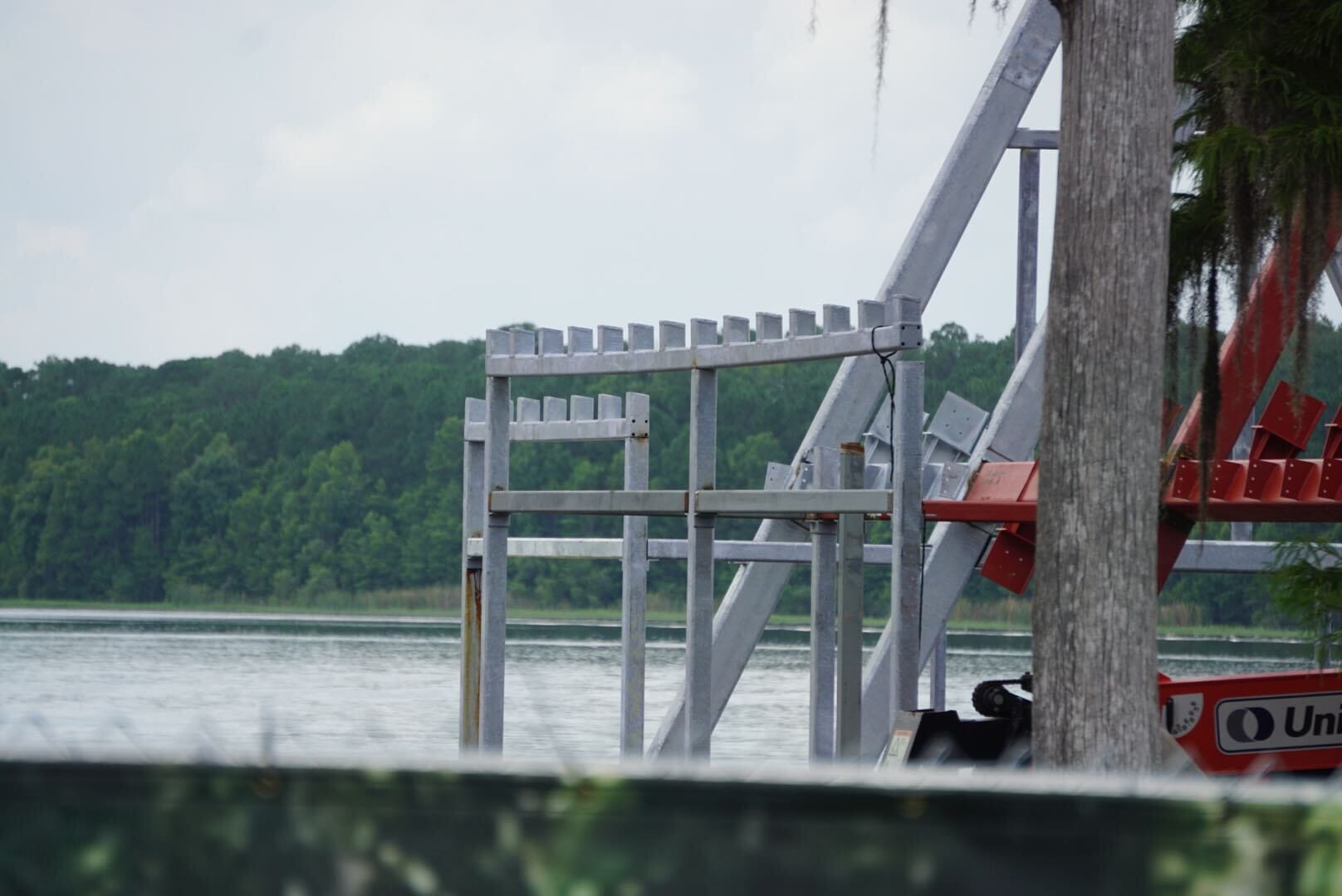 Metal scaffolding near a calm lake hints at ongoing lakeshore lodge construction under a cloudy sky and trees.