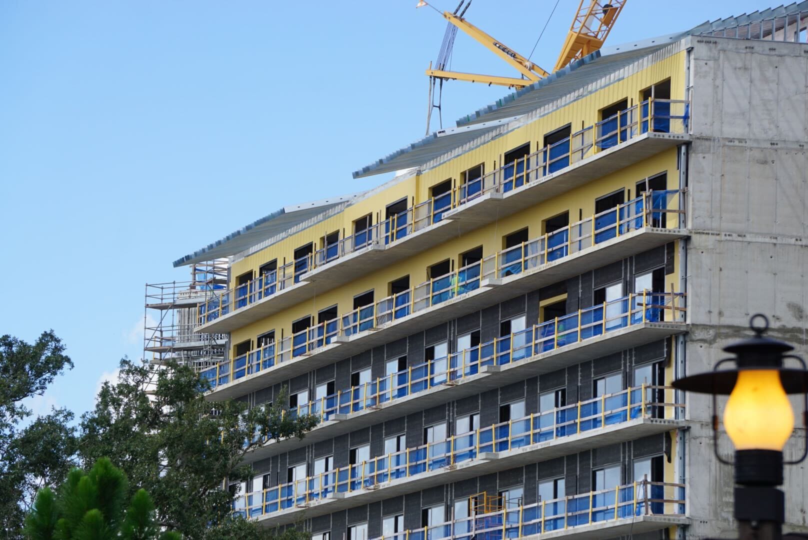 A multi-story building under construction with yellow panels and blue railings, cranes visible above, and a tree in front.