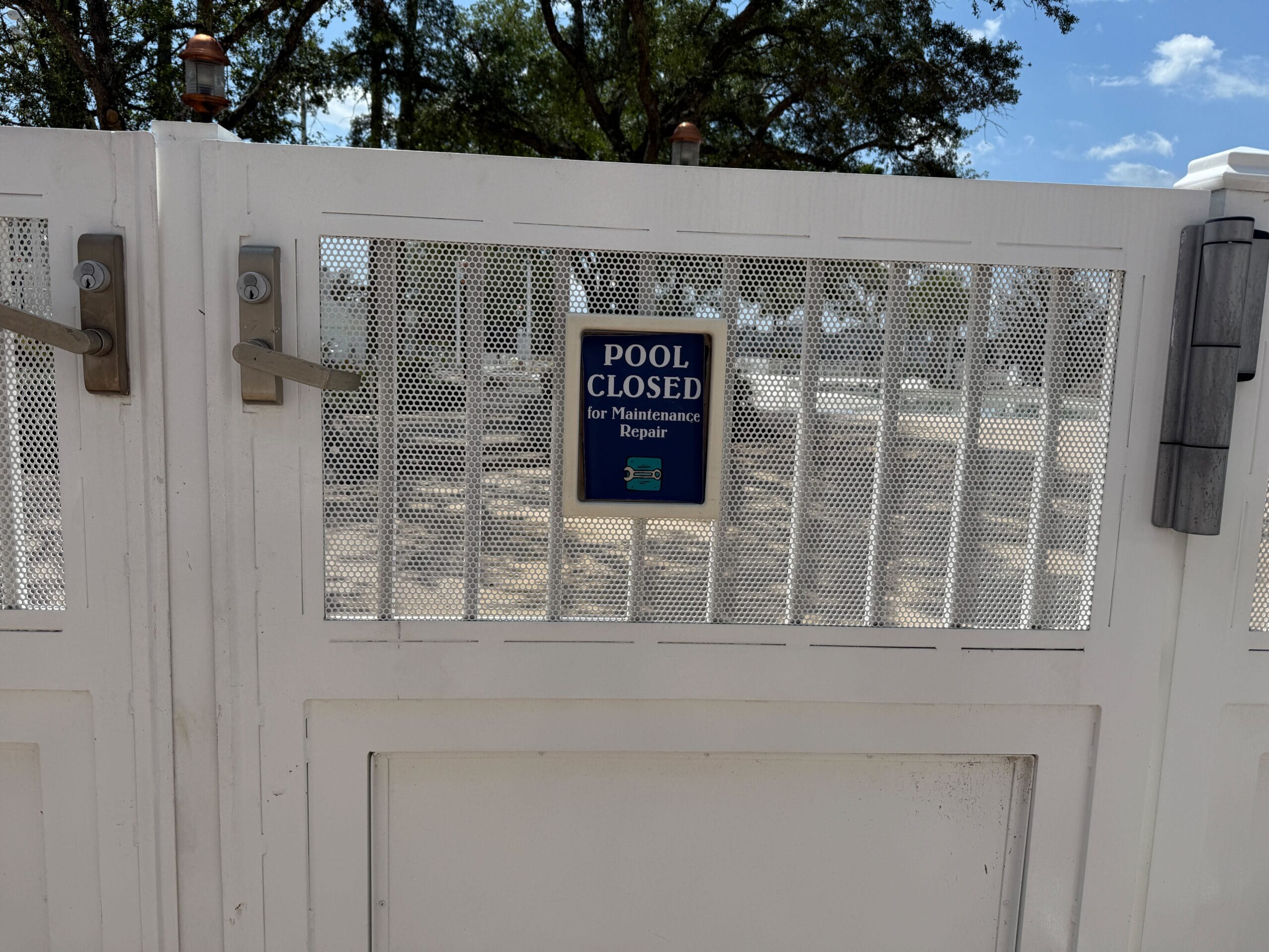 White metal gate with a sign that reads Pool Closed for Maintenance Repair in front of a sunny outdoor area.