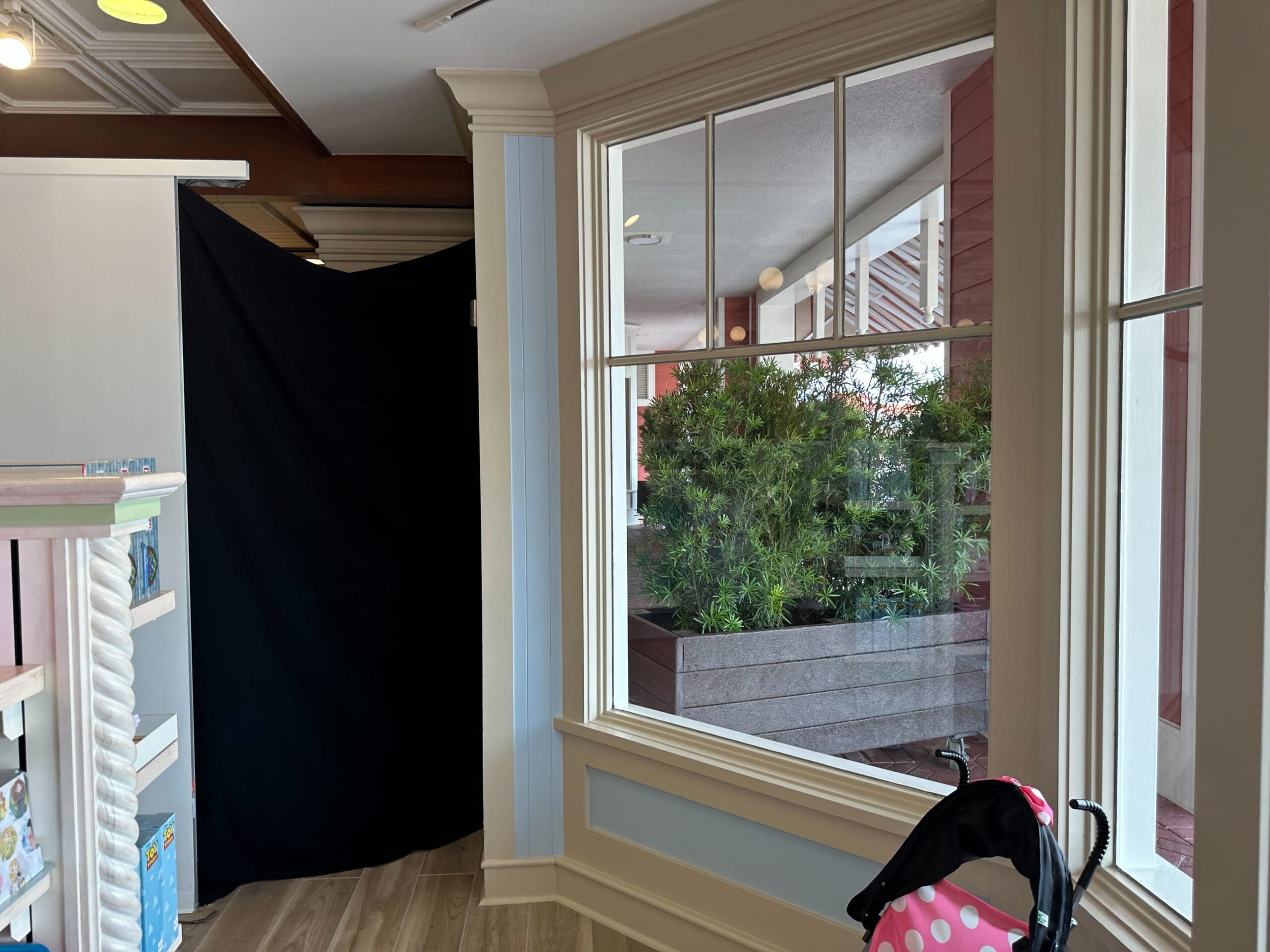 Interior view of a retail space, featuring the renovated section of Disney's Boardwalk Inn; through the large window, lush plants are visible outside alongside a partially visible stroller with pink polka dot fabric in the foreground.