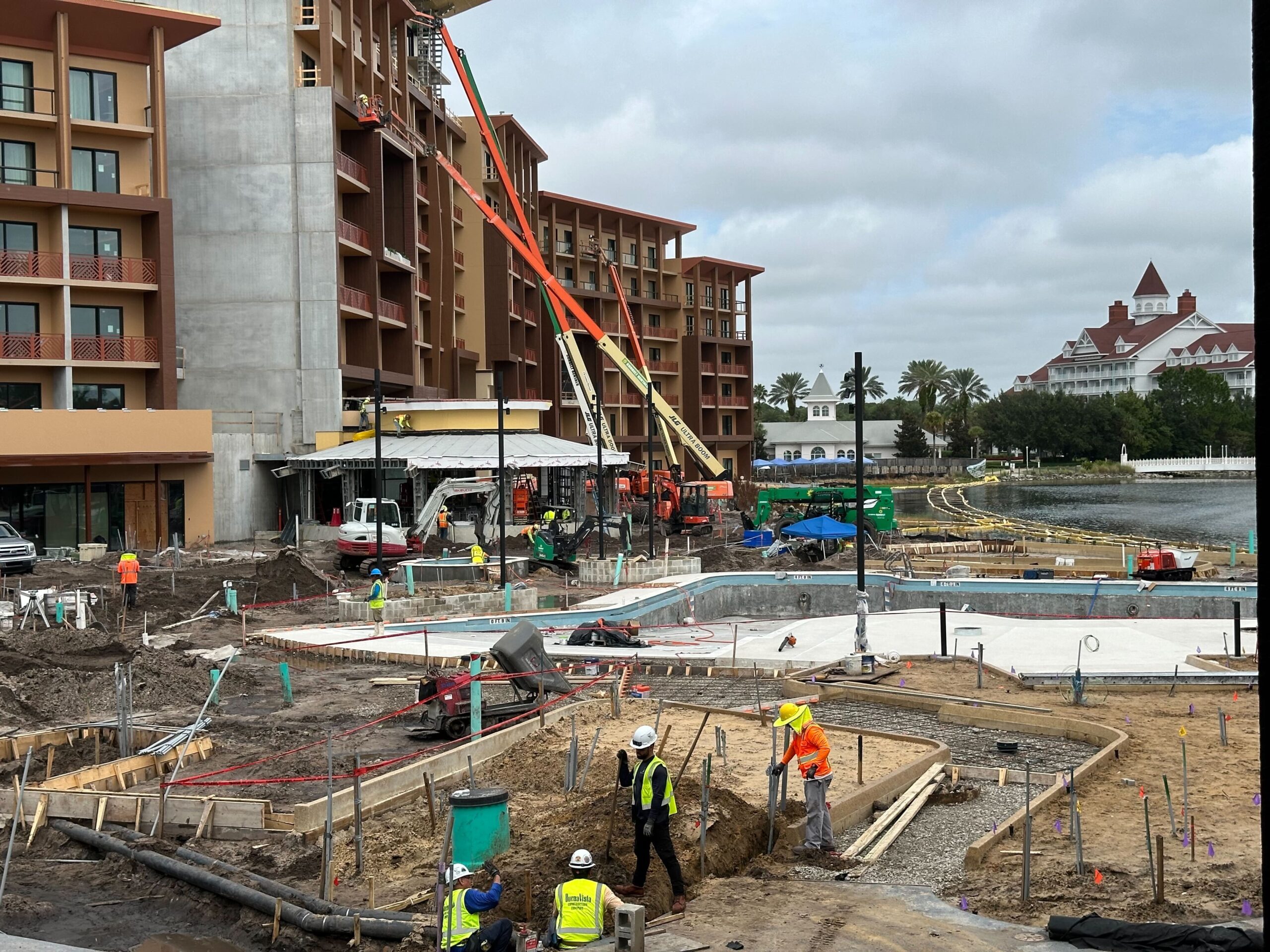 Construction site of a large building with workers operating machinery and performing various tasks. The site features heavy equipment, scaffolding, and partially completed structures in a waterfront area.