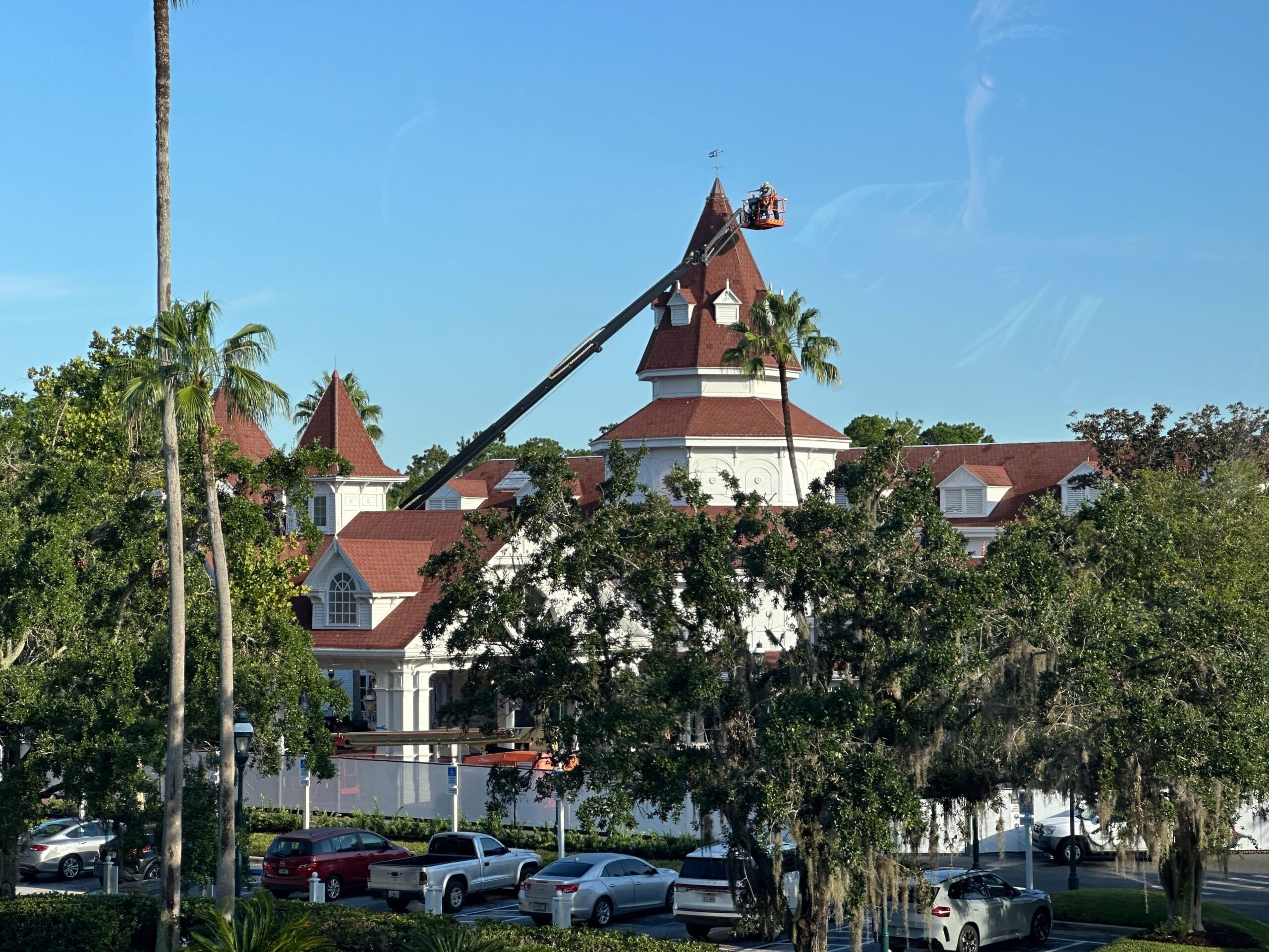 A crane lifts workers to the roof of a Grand Floridian-style building with red shingles, white walls, and surrounding trees.