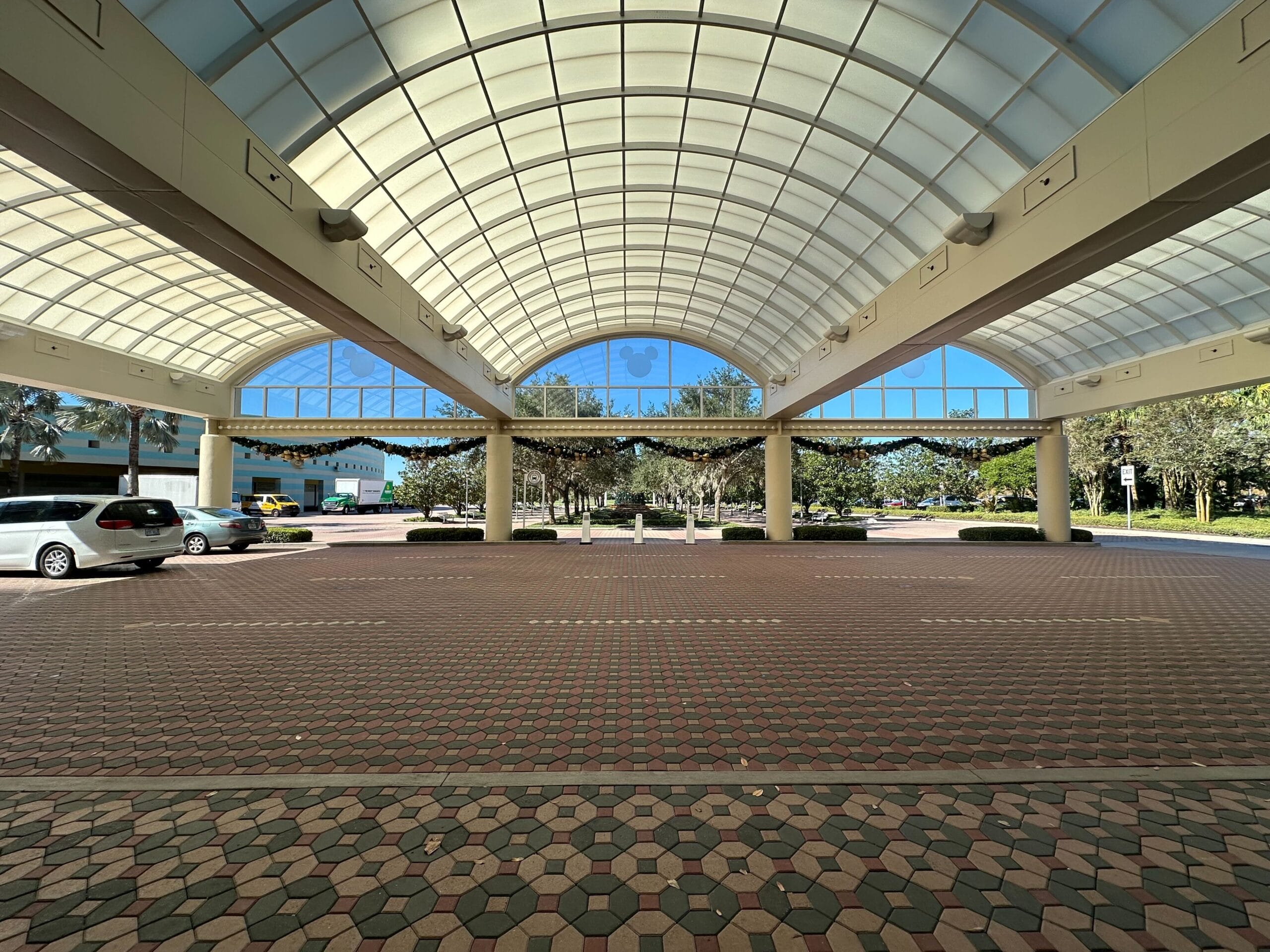 Spacious covered entrance with a curved glass roof and a hexagonal tile floor, surrounded by greenery and parked vehicles.