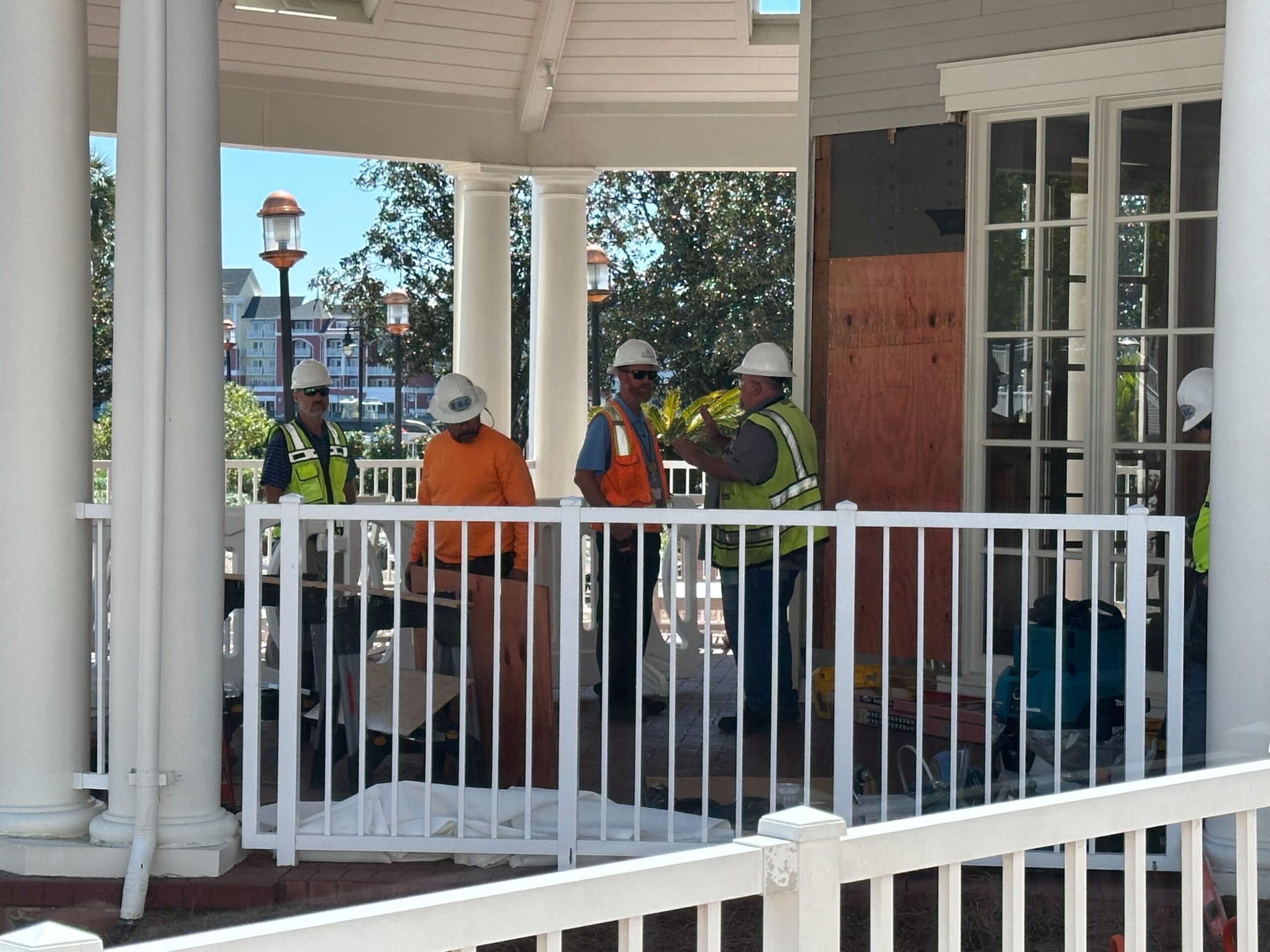 Construction workers in safety gear stand and work inside a fenced area of a white gazebo under a clear blue sky.