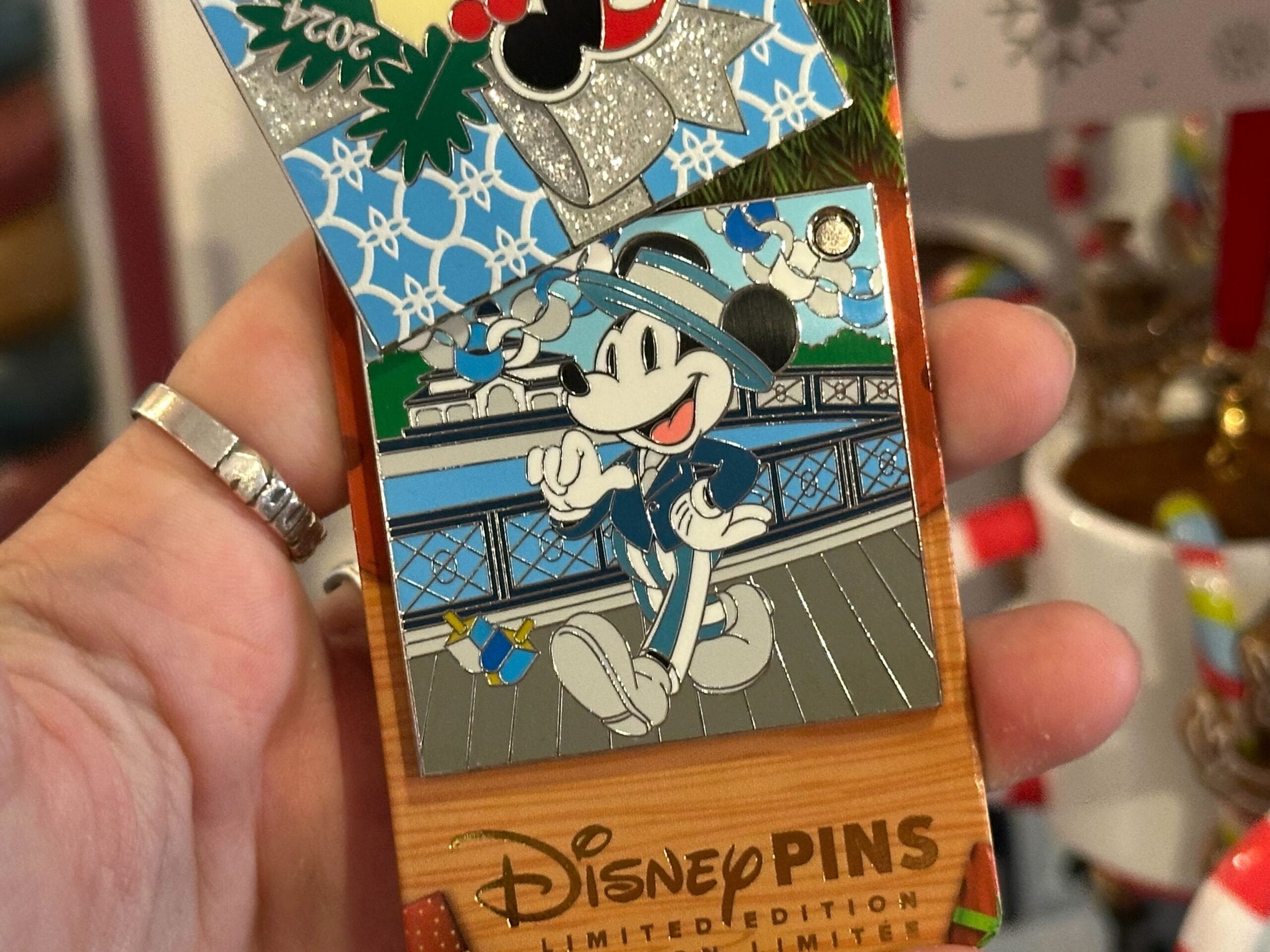 A hand holds a Disney Pins limited edition pin featuring a festive design with a character dressed in sailor attire, set against a waterfront background.