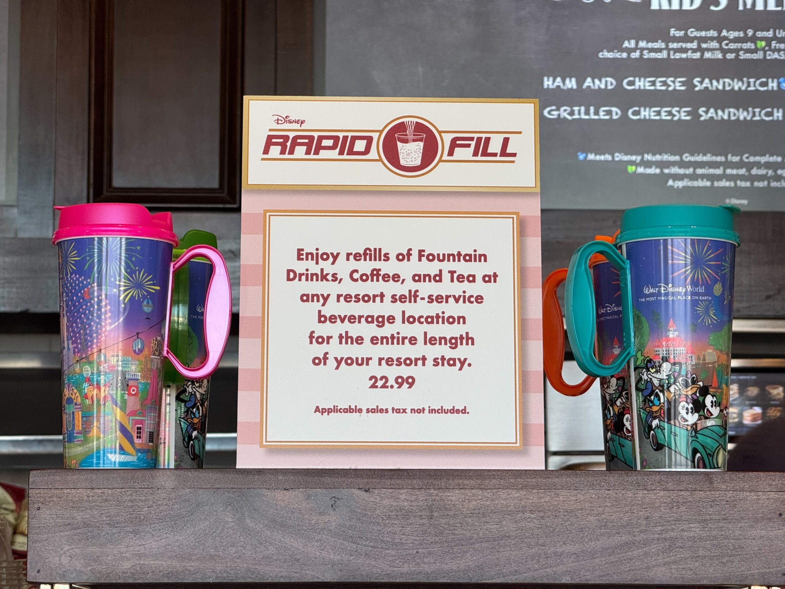 Two Resort Refillable Mugs with lids and handles sit on a counter beside a sign at Walt Disney World, offering unlimited beverage refills for $22.99 during your resort stay.