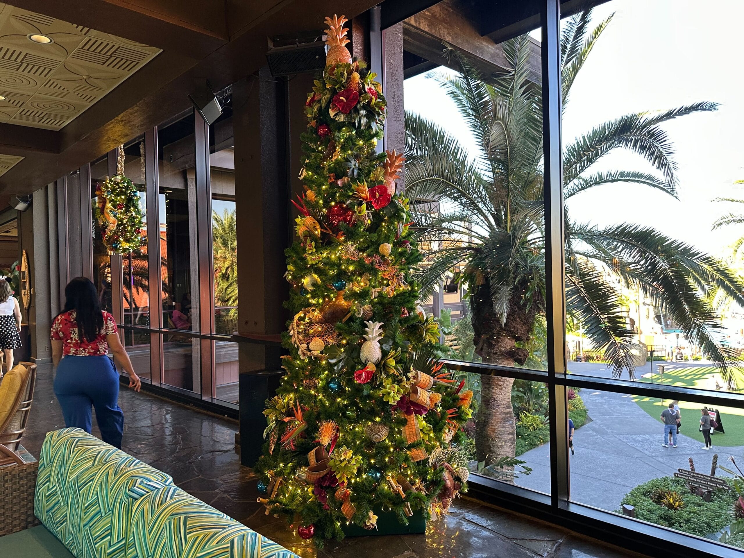 A decorated Christmas tree with lights stands inside a building next to large windows overlooking palm trees outside.
