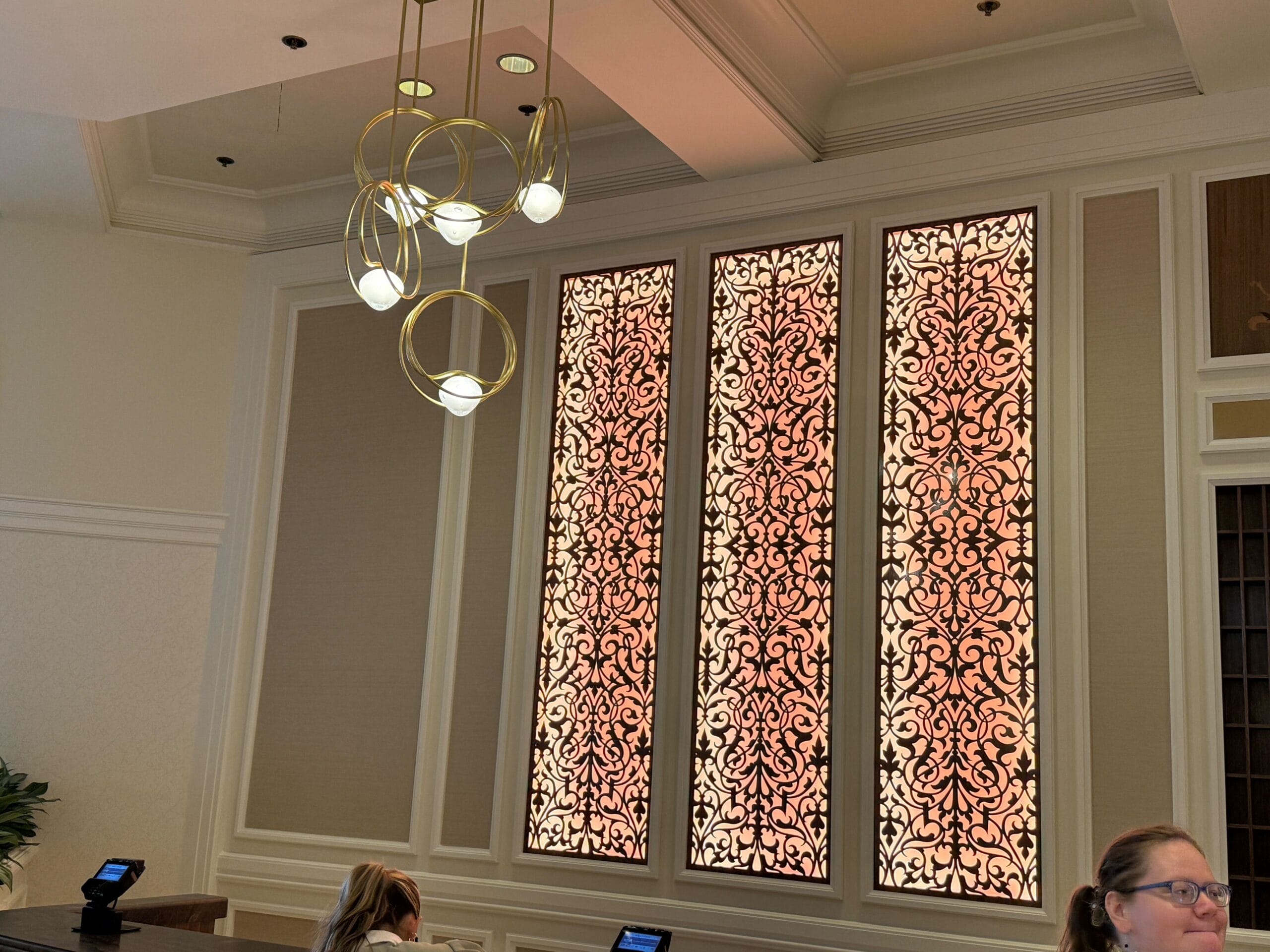Elegant lobby area with ornate backlit wall panels, modern gold chandelier, and two people at a counter.