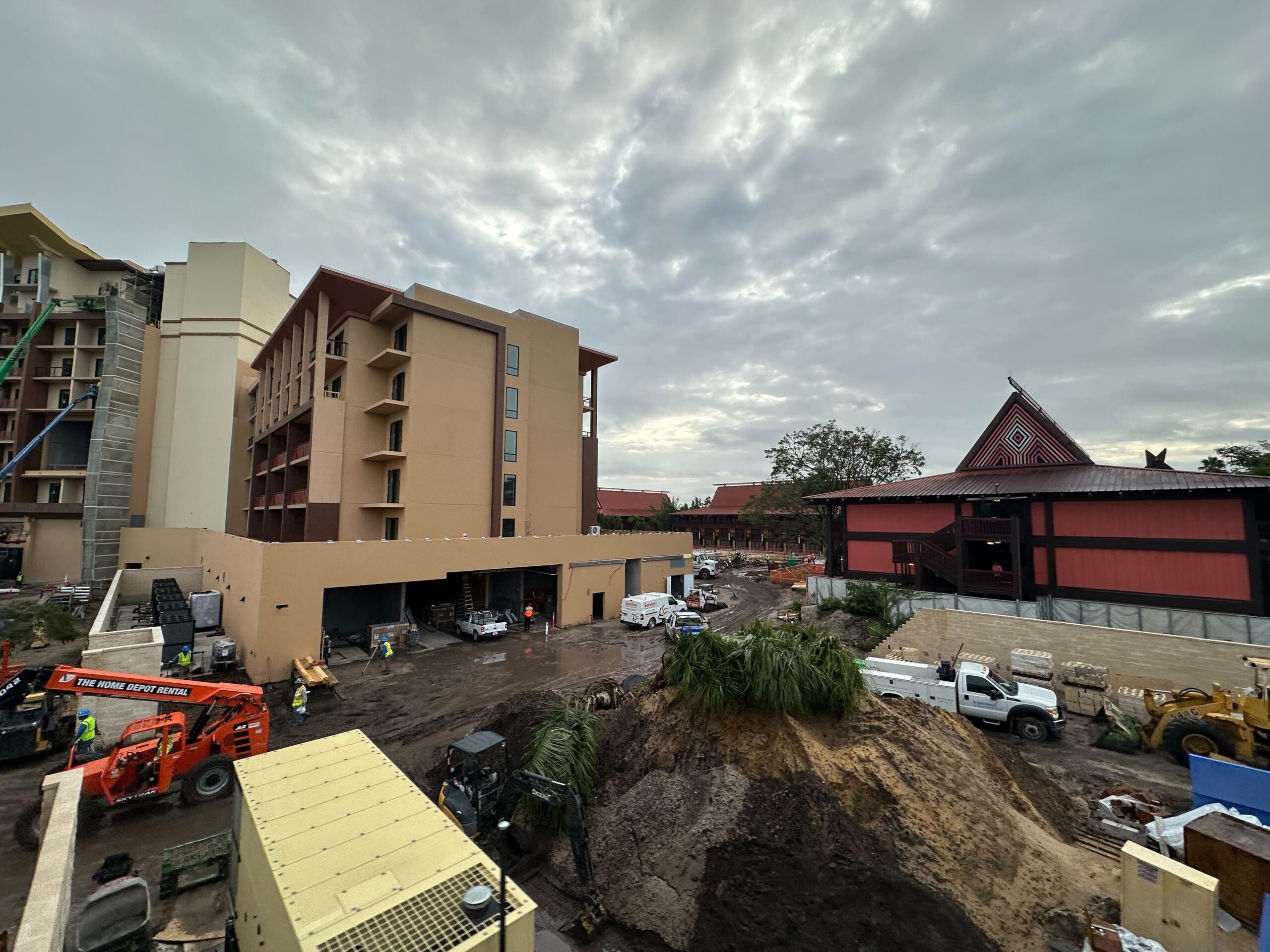 Construction site with several vehicles and equipment near multistory buildings and a pile of dirt. The sky is overcast.