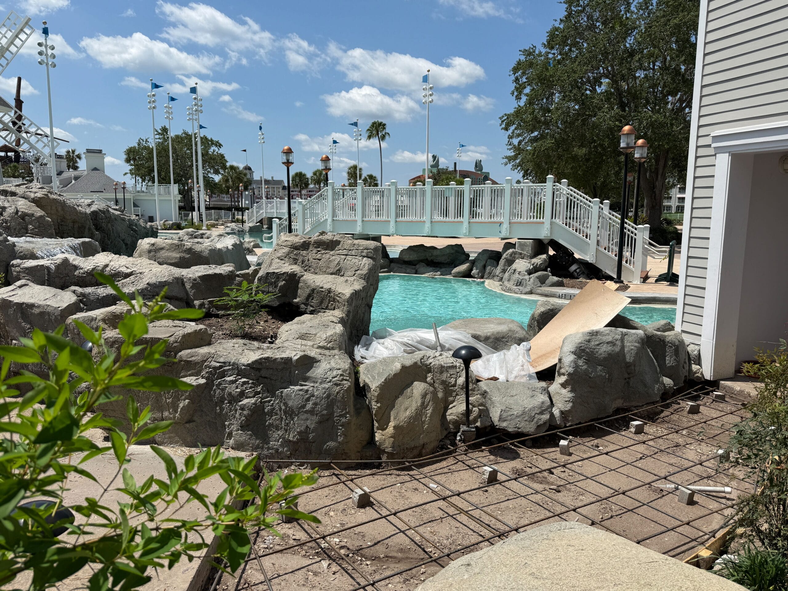 Rocky pool area under construction at Stormalong Bay, featuring a footbridge, greenery, and blue sky with scattered clouds.