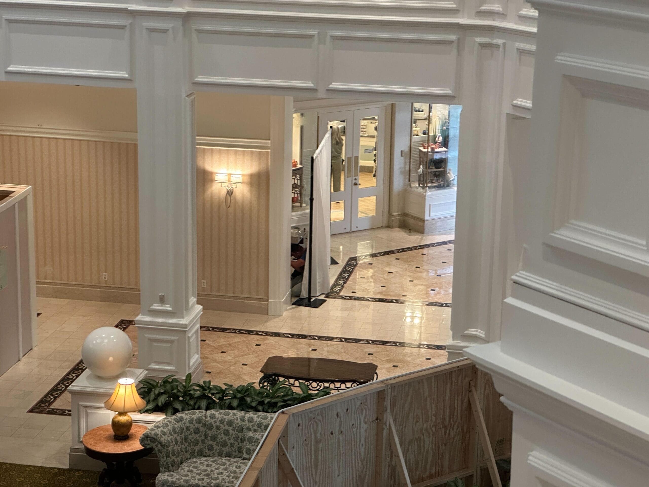 A hotel lobby with white walls, patterned chairs, a table lamp, and marble floors seen from a higher level.