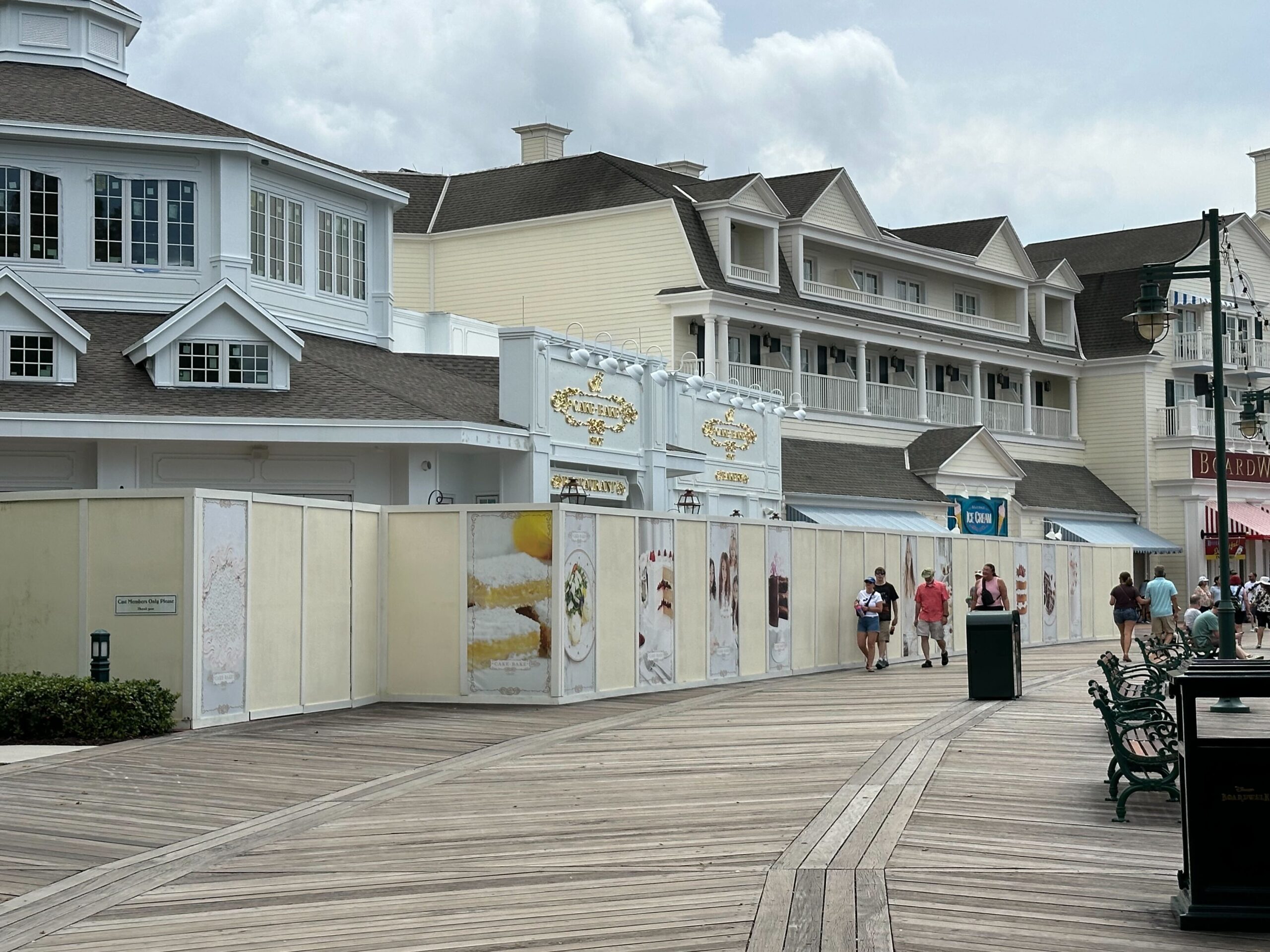 A boardwalk with construction walls featuring artwork and signage. Several people walk along the boardwalk in front of large, colonial-style buildings under a cloudy sky.