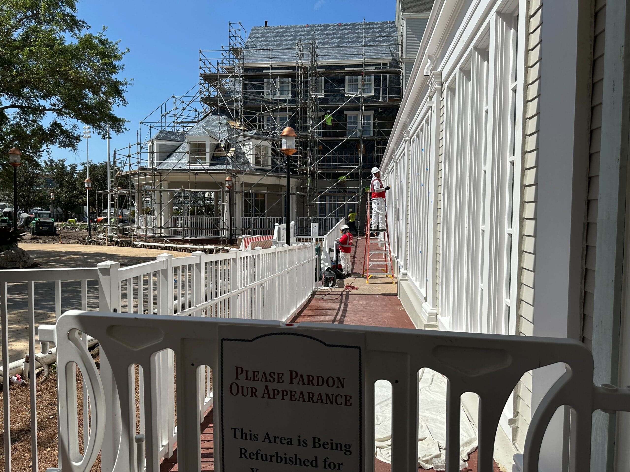The building, resembling Disney’s Yacht Club, is under renovation with scaffolding. Workers in protective gear are busy repainting the white fence. A sign kindly requests pardon for the temporary appearance during this enhancement phase.