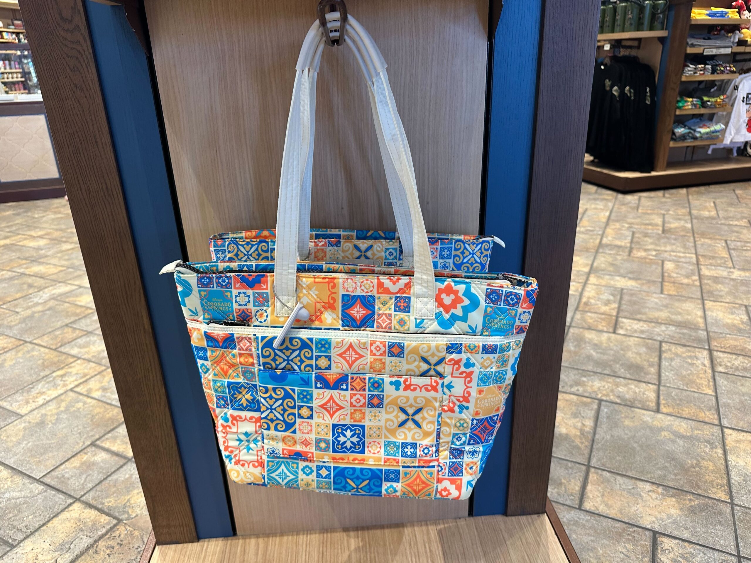 A multicolored Lug Bag with a tiled pattern hangs on a wooden display, showcasing its vibrant charm amidst the Coronado Springs Merchandise collection in the store.