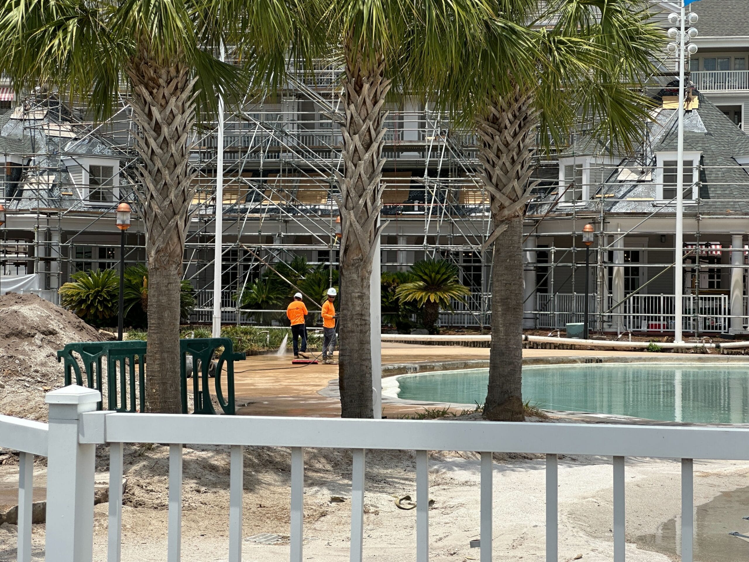 Two construction workers in orange vests stand near scaffolding by a pool, with palm trees and a white fence in front.