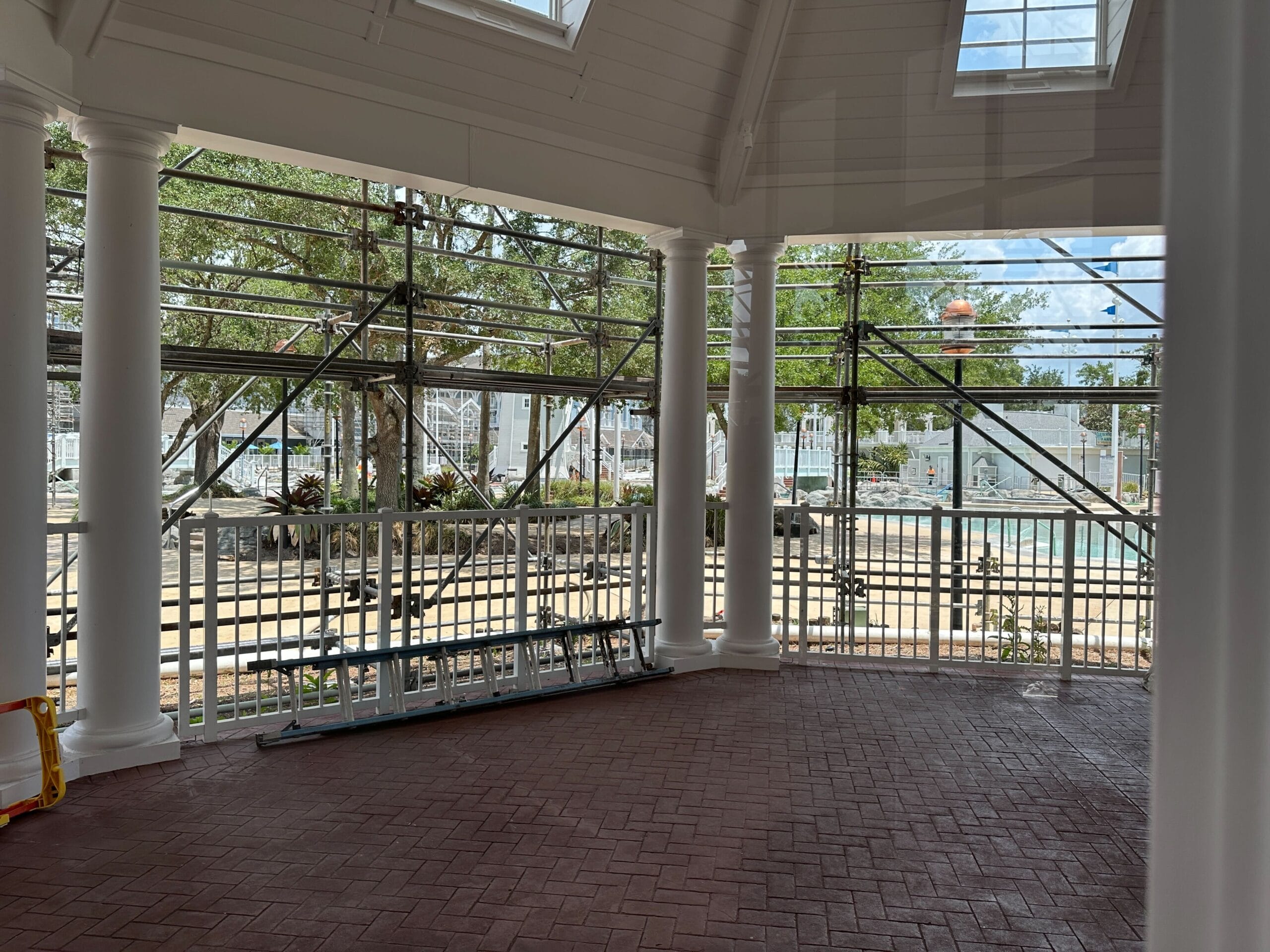 Covered patio with white columns, scaffolding, and a ladder, overlooking a pool and trees outside.