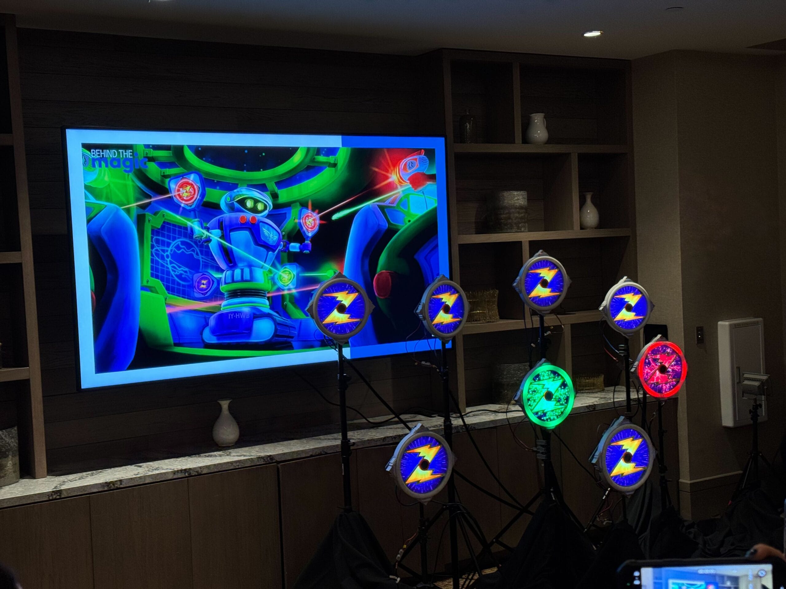 A brightly lit room features a large screen displaying a colorful cartoon robot. Several circular lights with lightning bolt designs are positioned in front of the screen.
