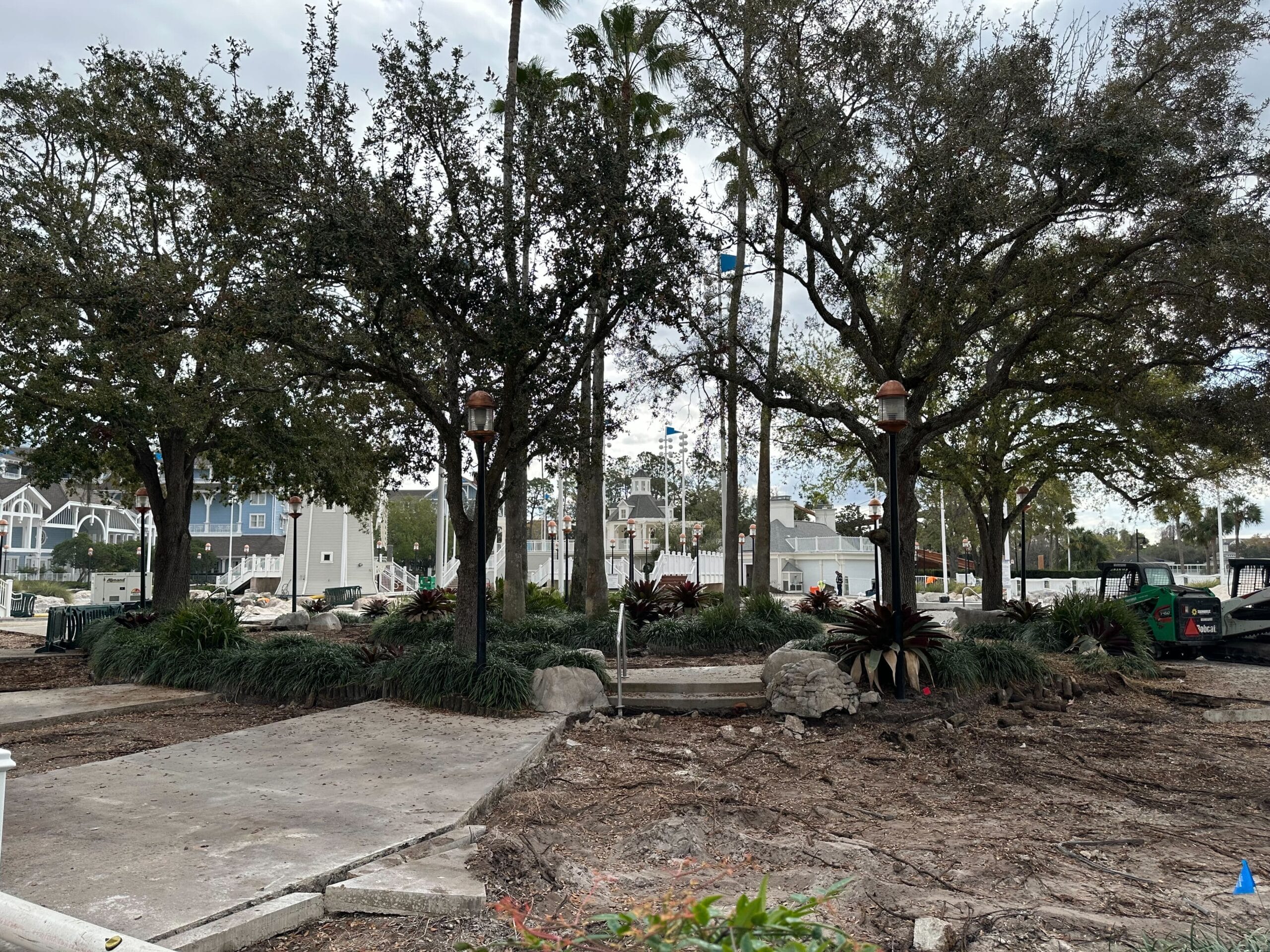Urban park under renovation with tall trees, some construction debris, and a fenced area in the background.
