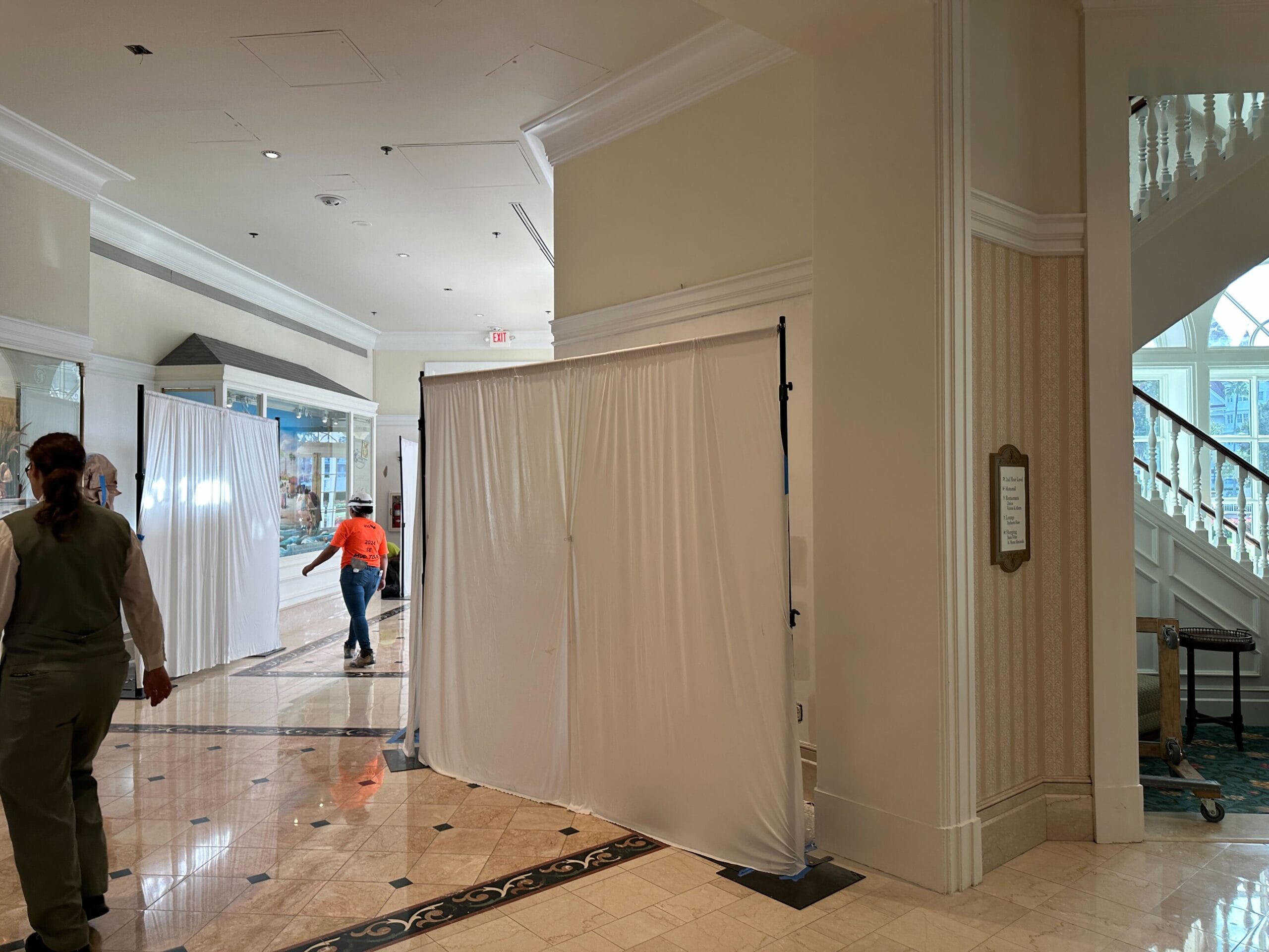 White sheets on stands block off part of a hotel lobby; two people walk near the construction area.