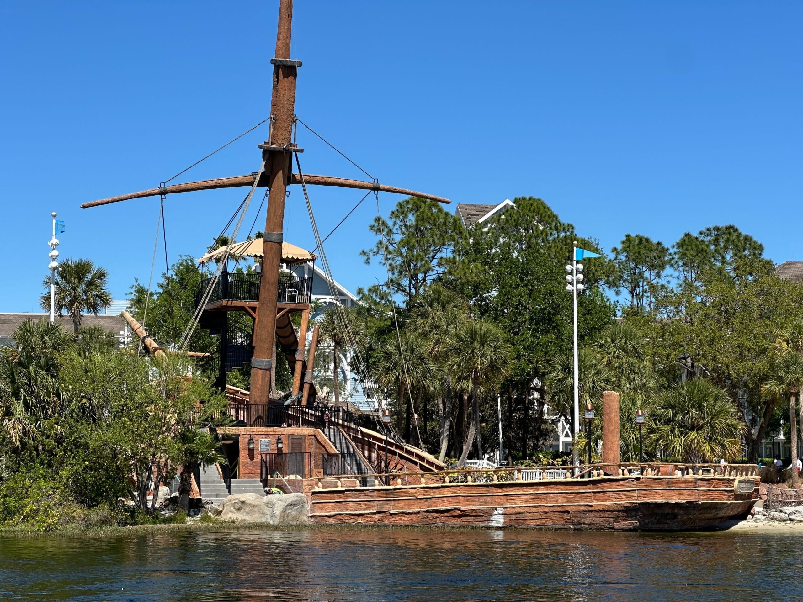 A wooden pirate ship with masts is docked by a lush, tree-lined shore under a clear blue sky, reminiscent of the enchanting ambiance at Disney’s Yacht Club.