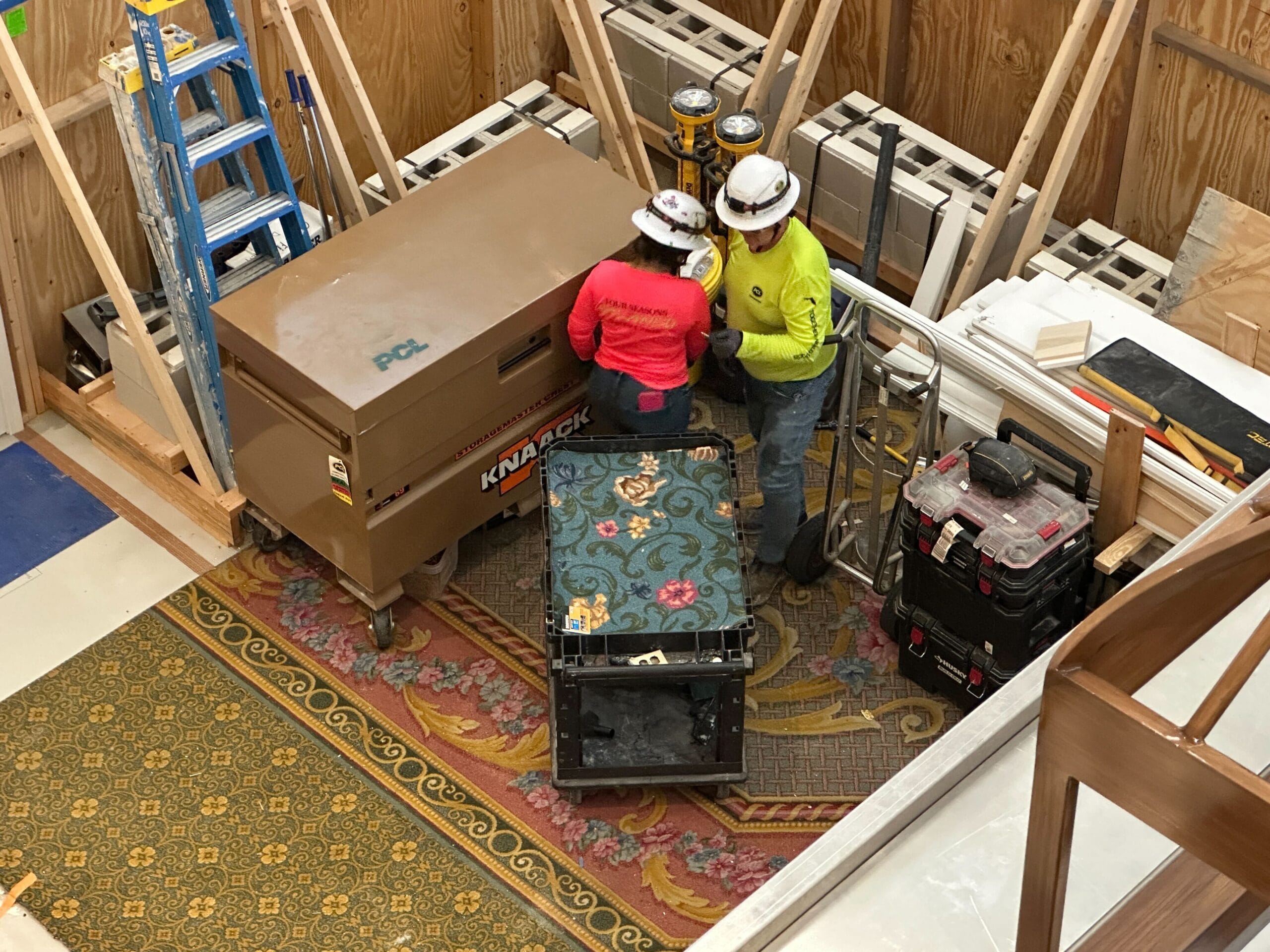 Two construction workers in safety gear stand by a toolbox and equipment in a wood-paneled room under construction.