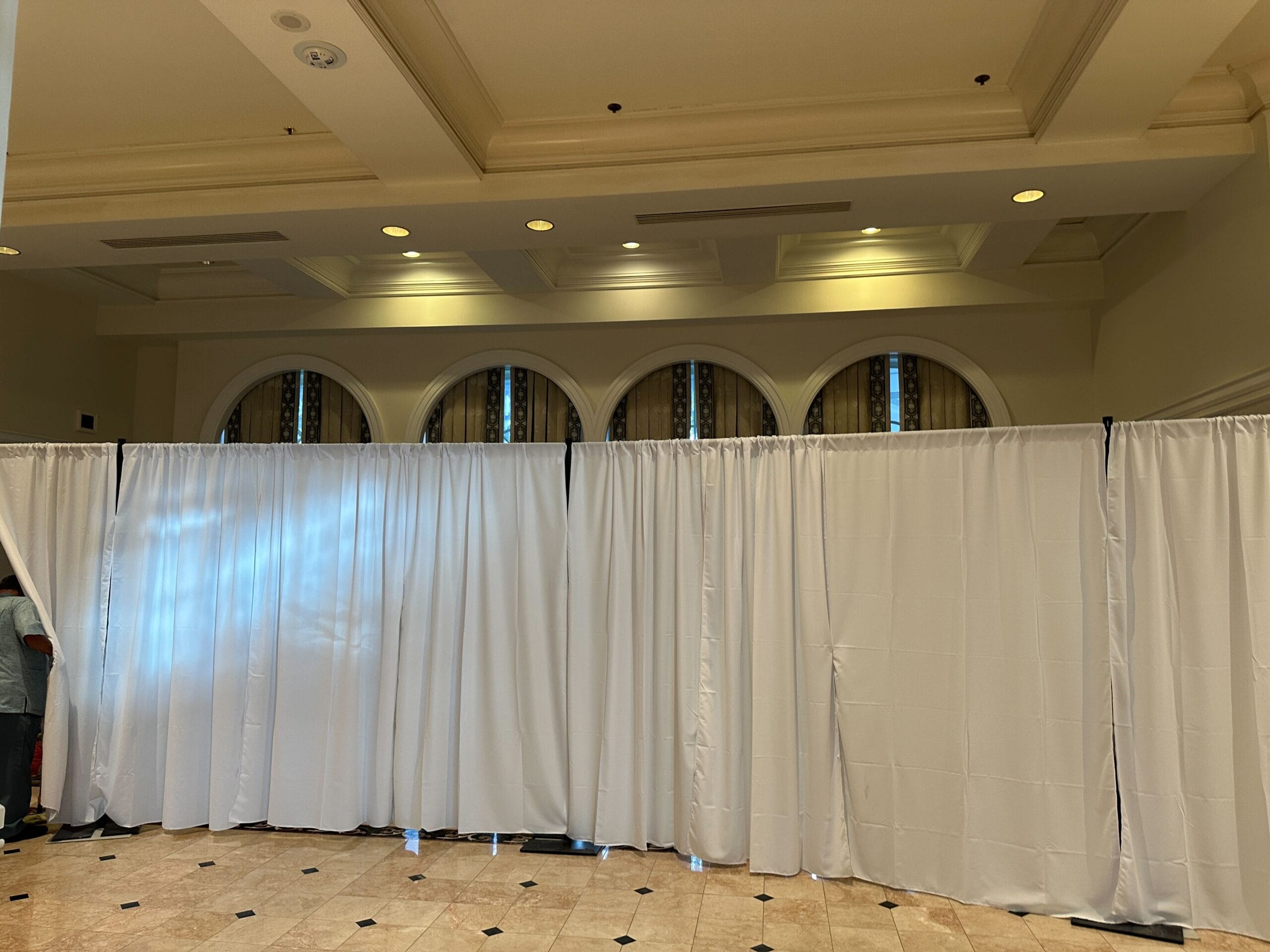 White curtains hang across a room, partially blocking arched windows and tiled floor in a well-lit indoor space.