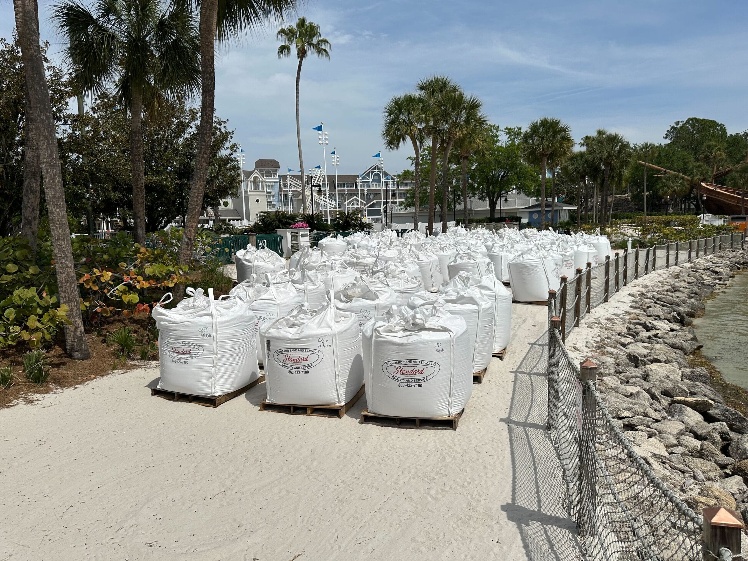 Large white sandbags are lined up on a sandy path near a body of water, suggesting pool construction in progress. Palm trees, a wire fence, and Disney's Yacht Club with flags fluttering in the background complete the picturesque scene.