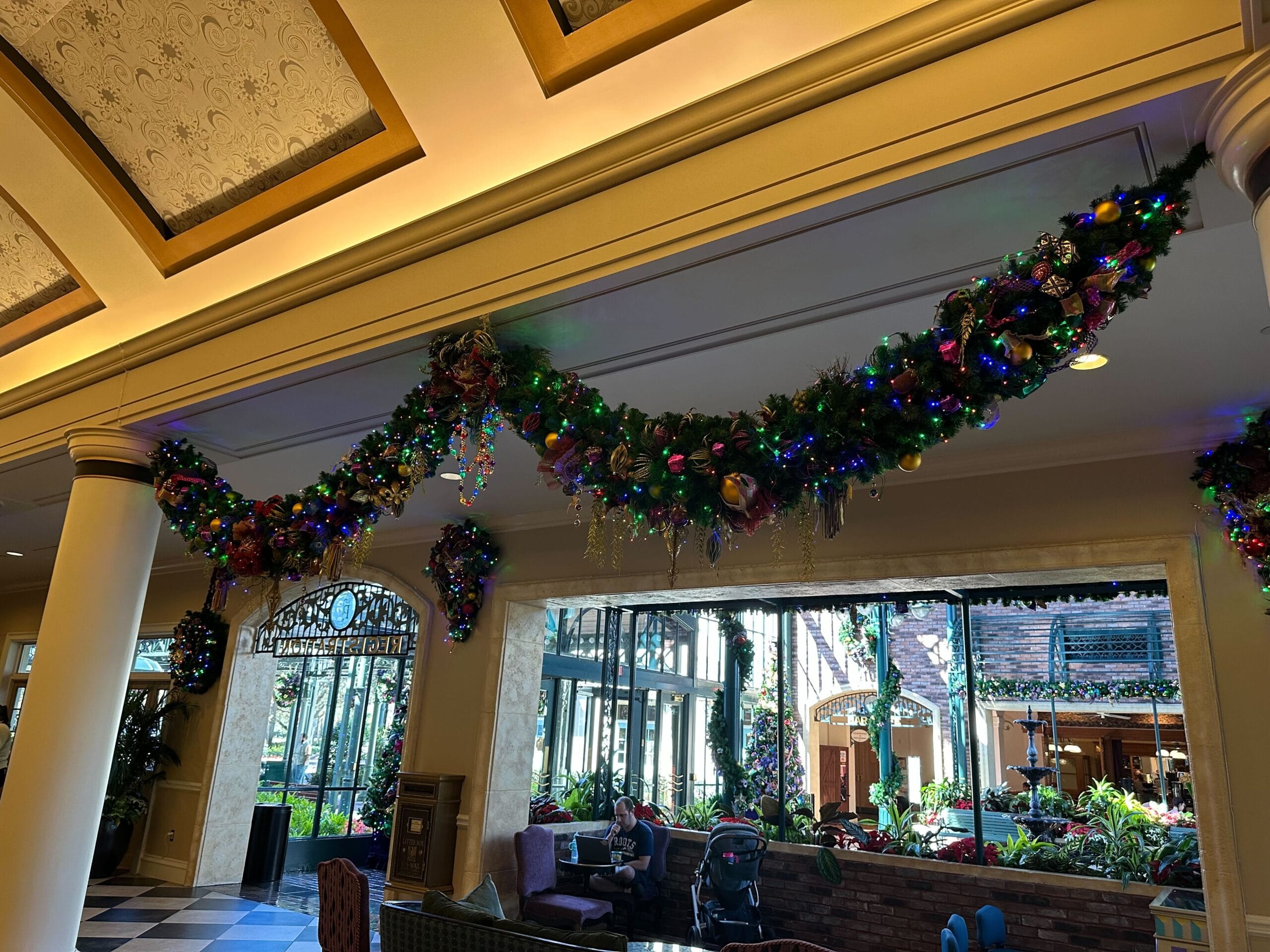 A decorated indoor space with garlands of greenery and multicolored lights hanging along the ceiling. Large windows reveal a garden outside.