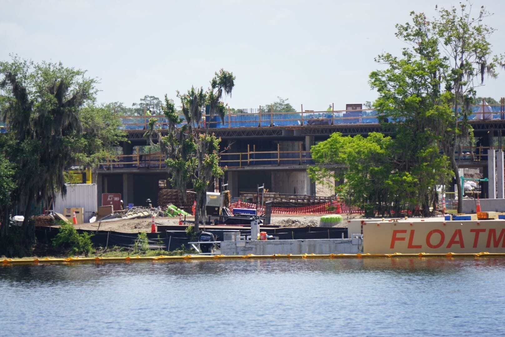 Construction site on the water with a partially built structure in the background, surrounded by trees and a barge labeled FLOATM in the foreground.